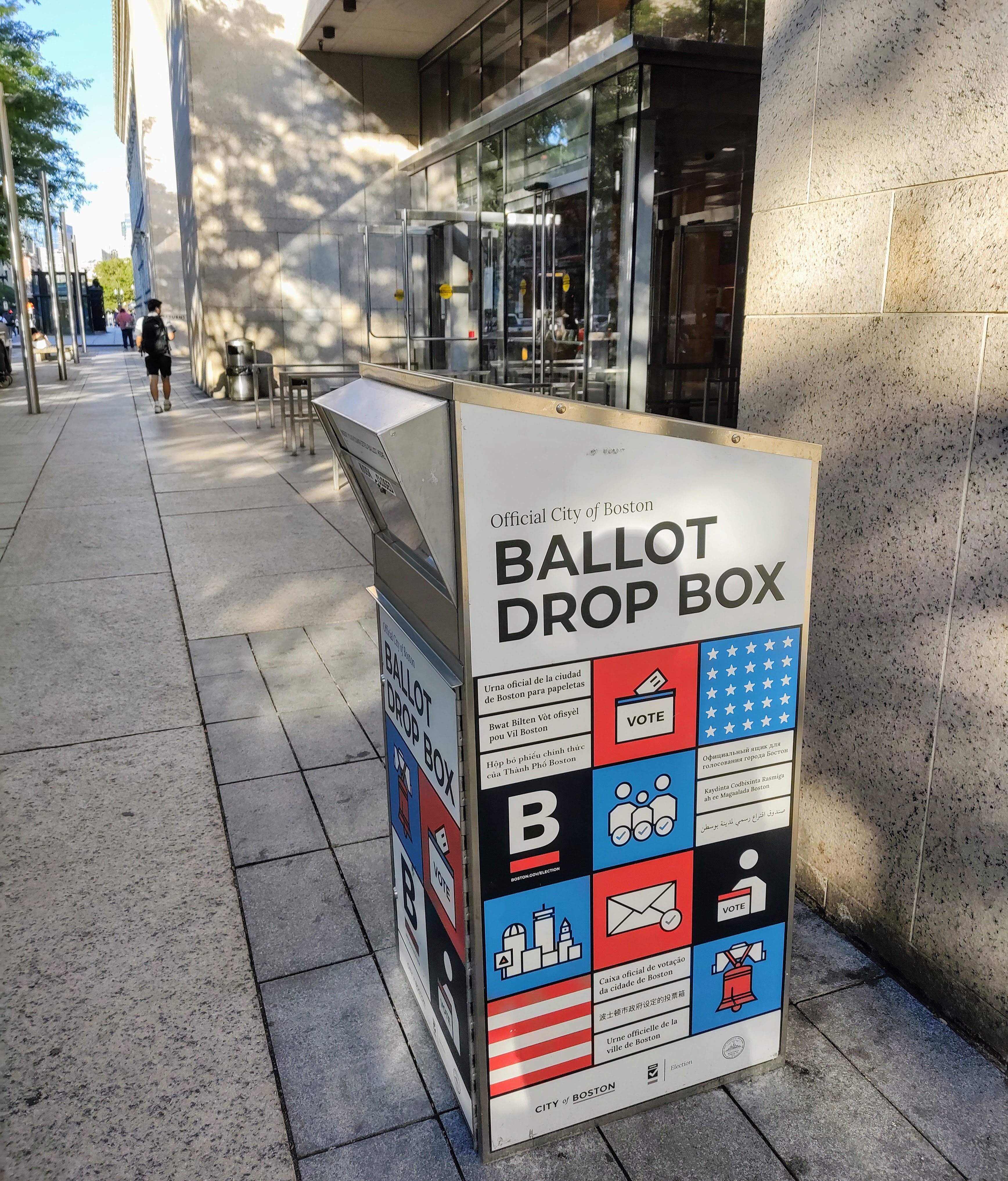 A red, white, and blue official ballot drop box on a sidewalk outside a building.