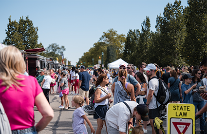 A large crowd walks along a tree-lined path at an outdoor festival on a sunny day.