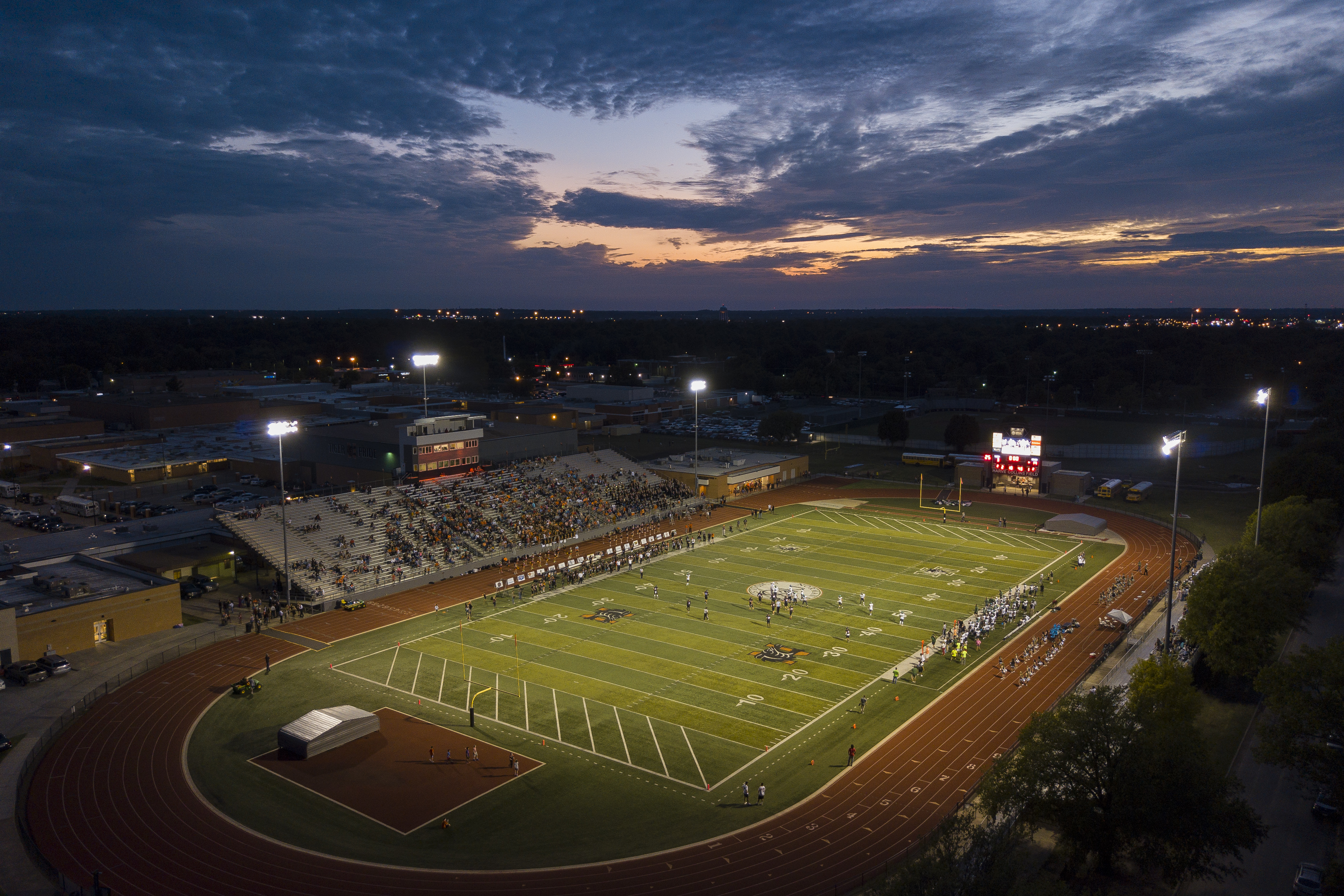 Aerial dusk view of a high school football field with stadium lights illuminated and spectators in the bleachers.