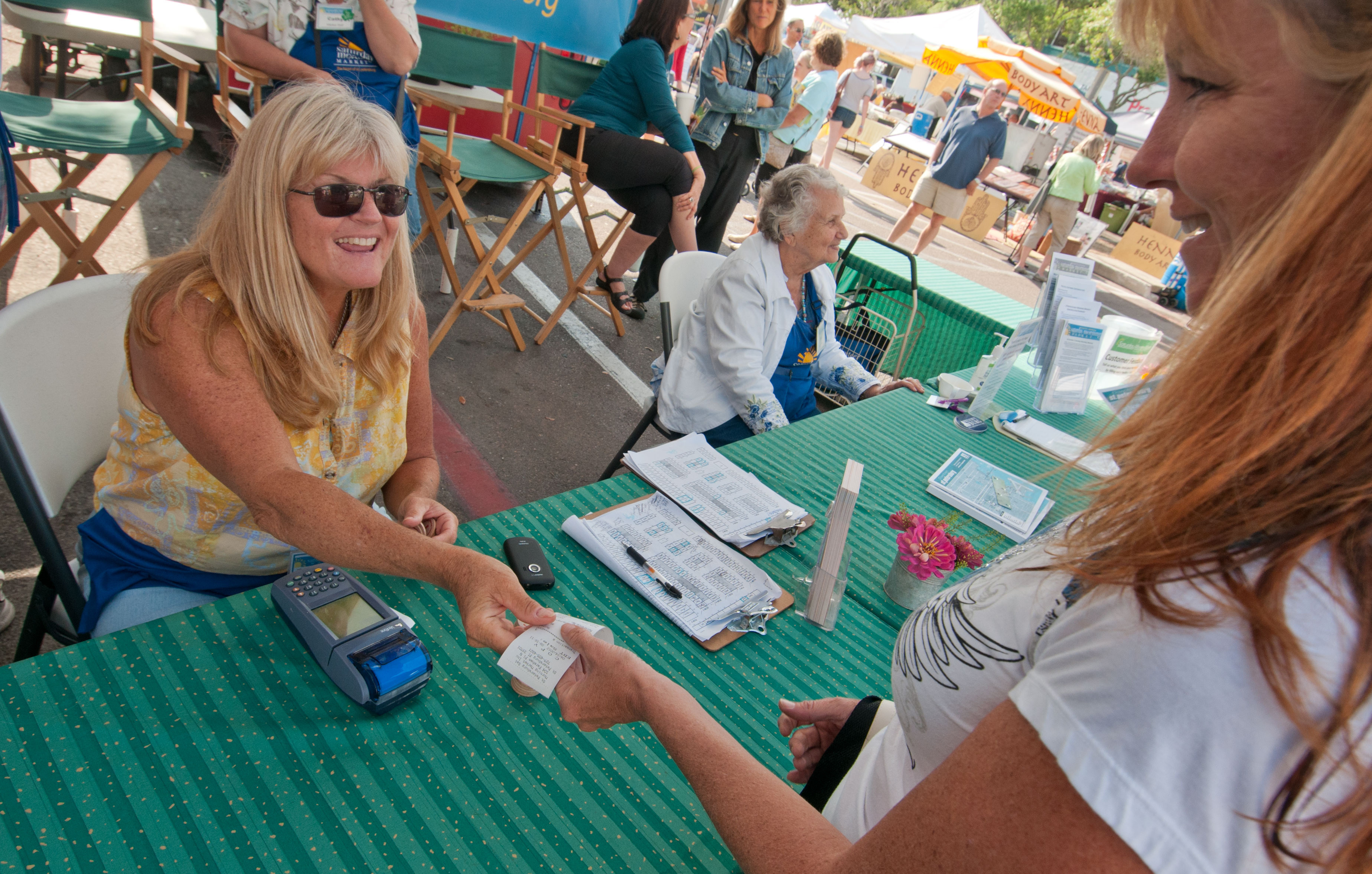 Two women completing a card transaction at an outdoor farmers market table.