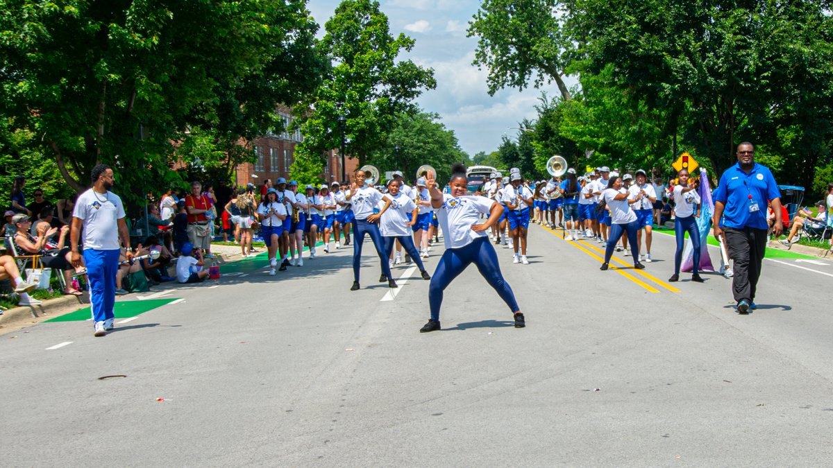 Marching band with dancers in white and blue uniforms performing on a tree-lined street with spectators on both sides.