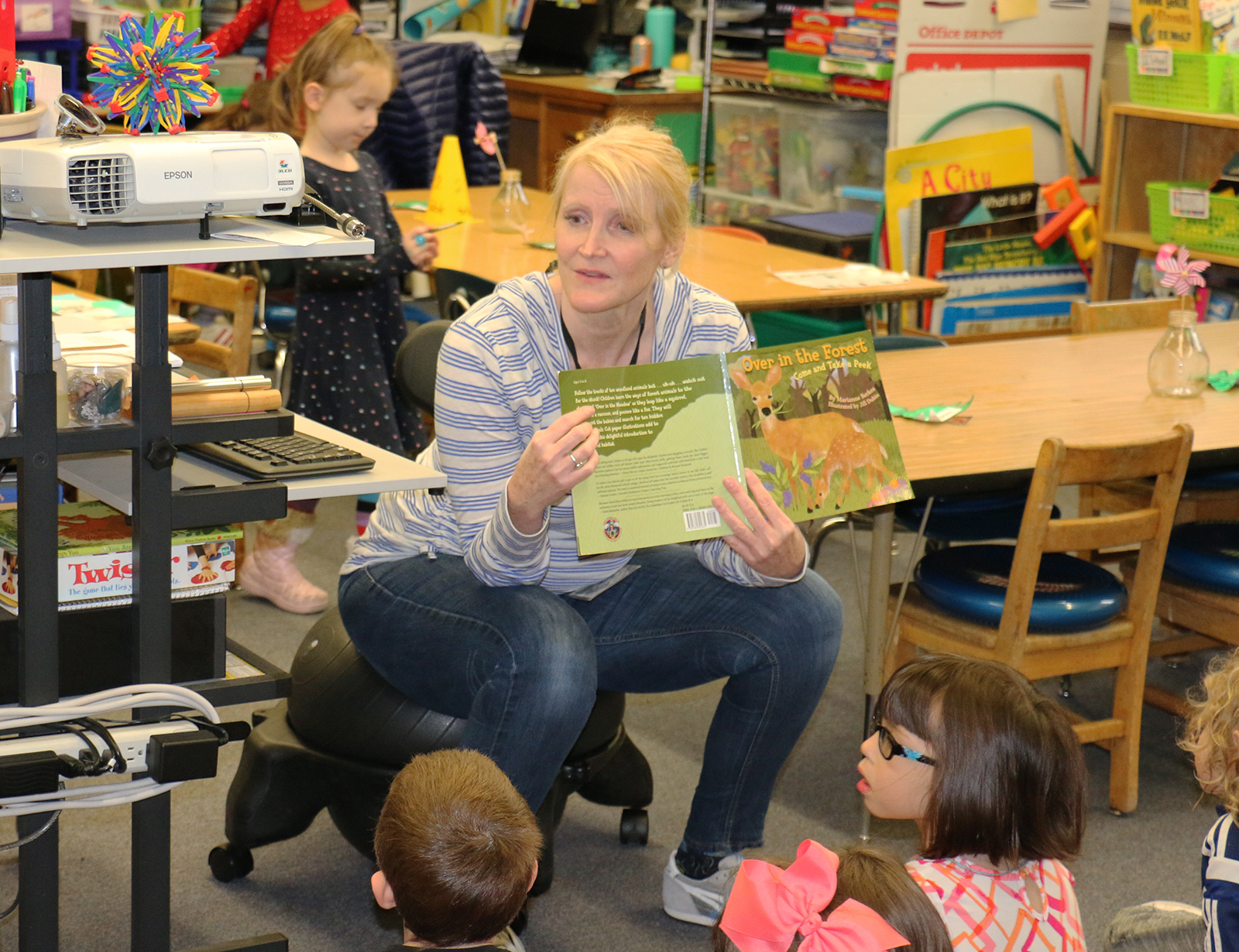 A woman reads a picture book aloud to young children seated on a classroom floor.
