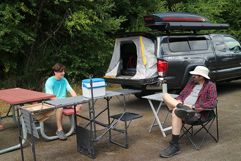 Two campers seated at a picnic table beside a pickup truck with a tent in the truck bed.