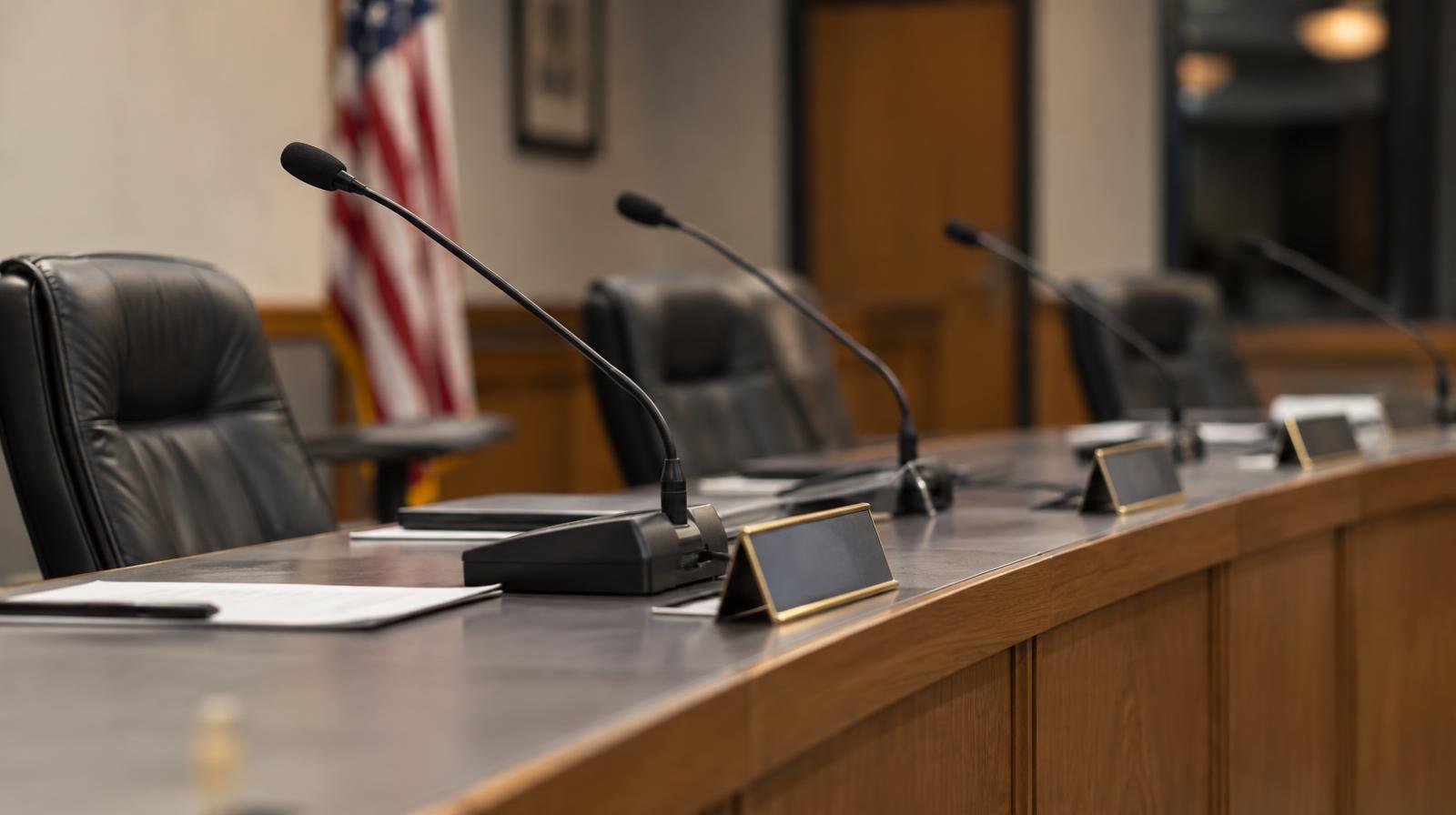 Empty board meeting room with a long dais, name placards, and microphones under institutional lighting.