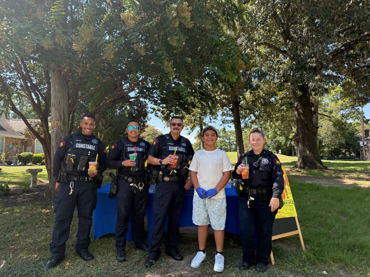 Four uniformed constable deputies and a child stand outdoors holding drinks near a blue table.