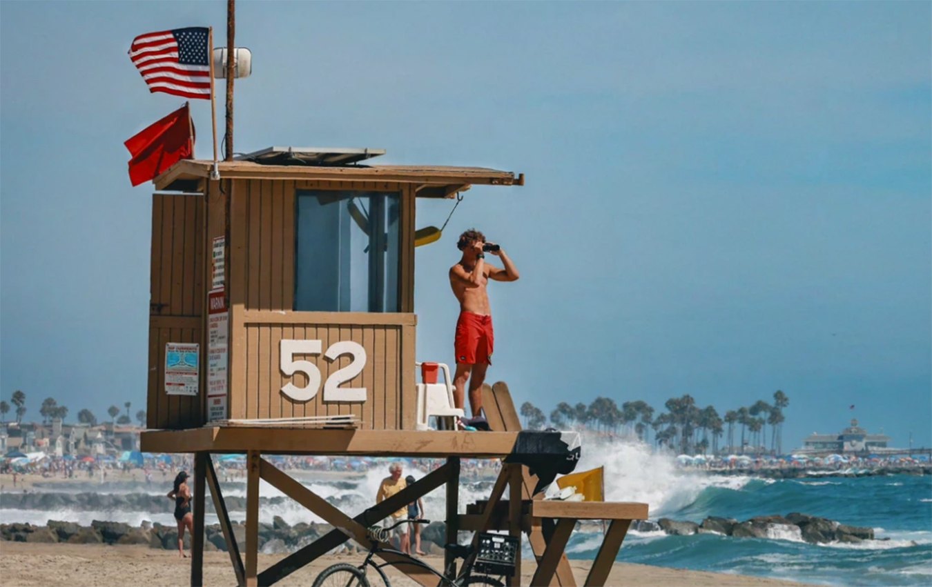 A lifeguard standing on an elevated wooden tower with a red warning flag and American flag flying above.