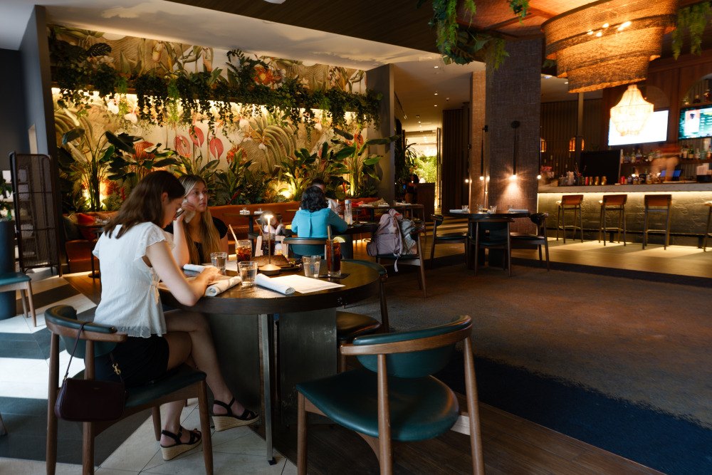 Restaurant interior with tropical plant-covered walls, a backlit bar, and wooden chairs.