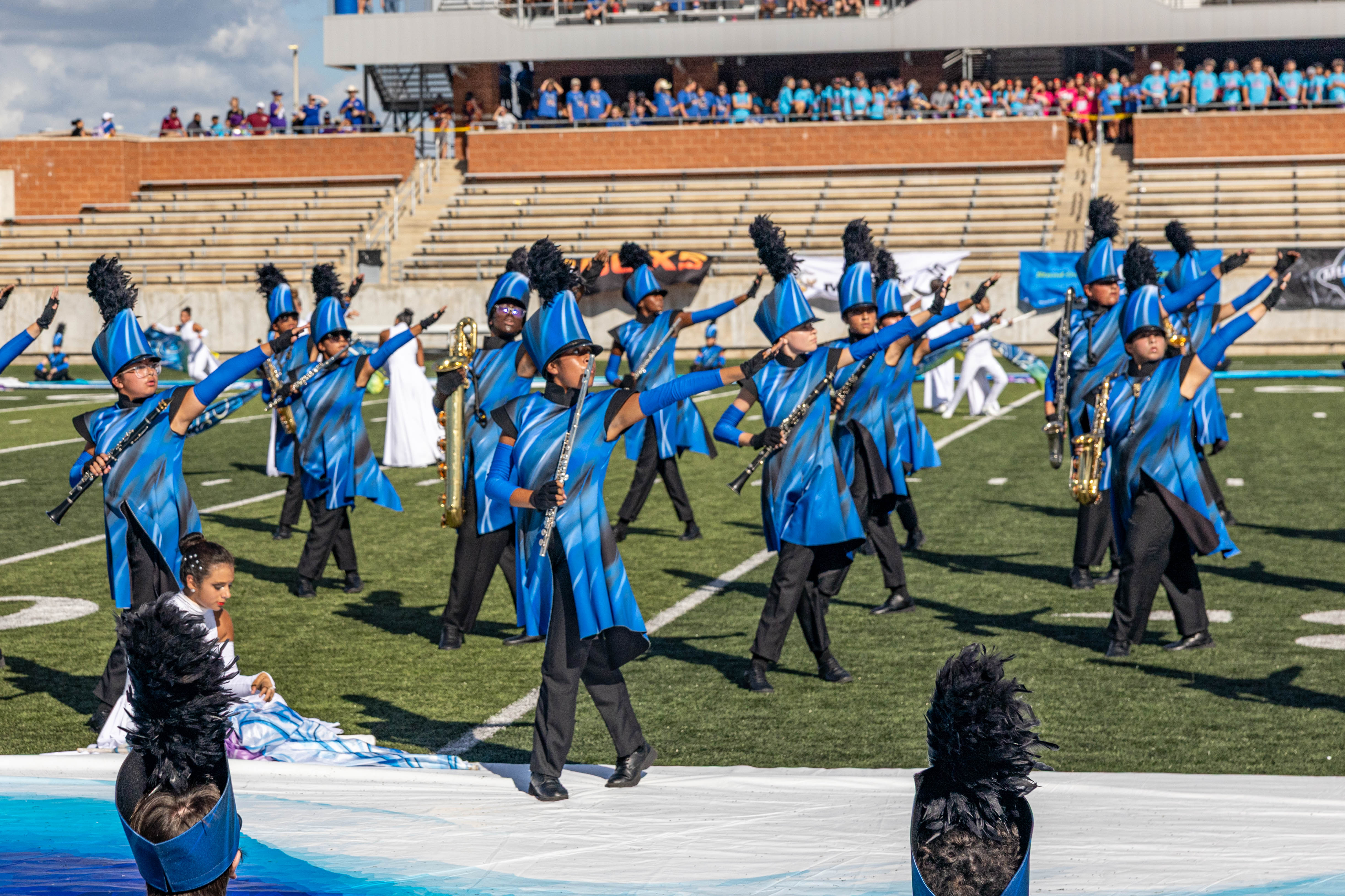 High school marching band in blue and black uniforms performing on a football field at a stadium.