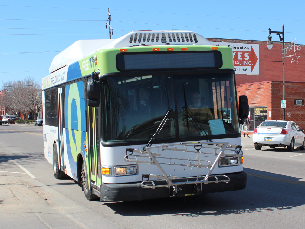 A green and white city transit bus with a front bike rack on a city street.