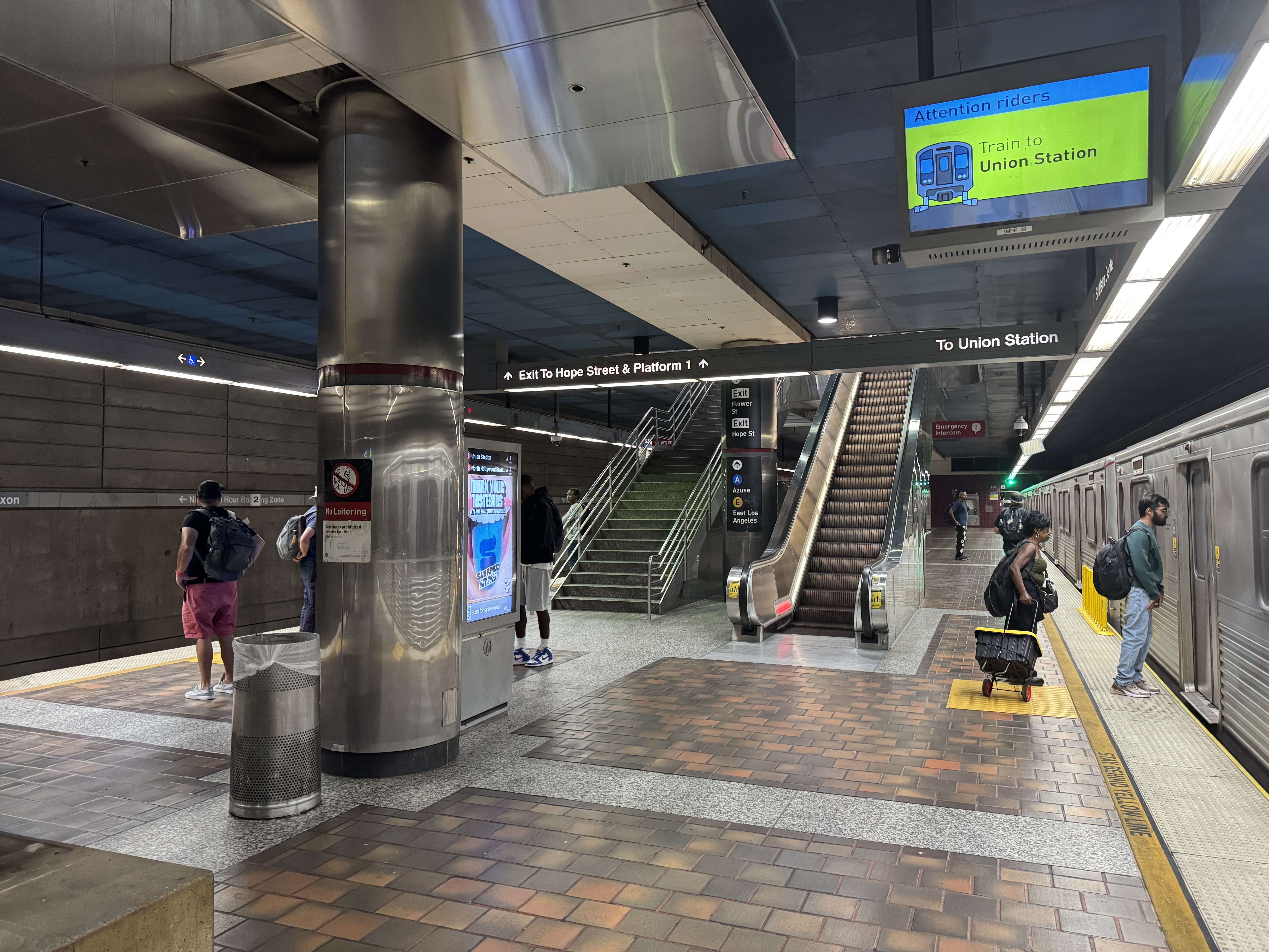 Passengers wait on an underground LA Metro platform with escalators and a train alongside.