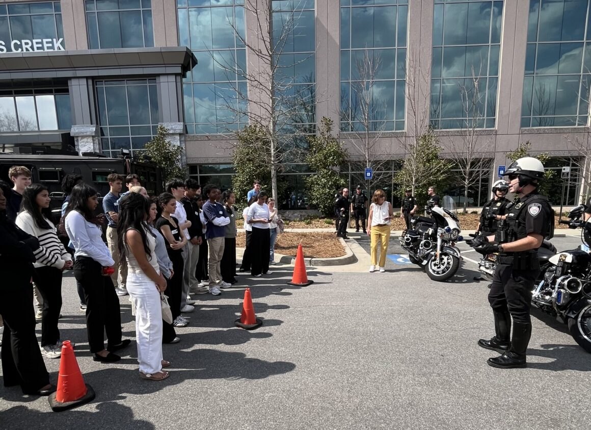 Two police officers on motorcycles address a group of students in a parking lot outside a modern office building.