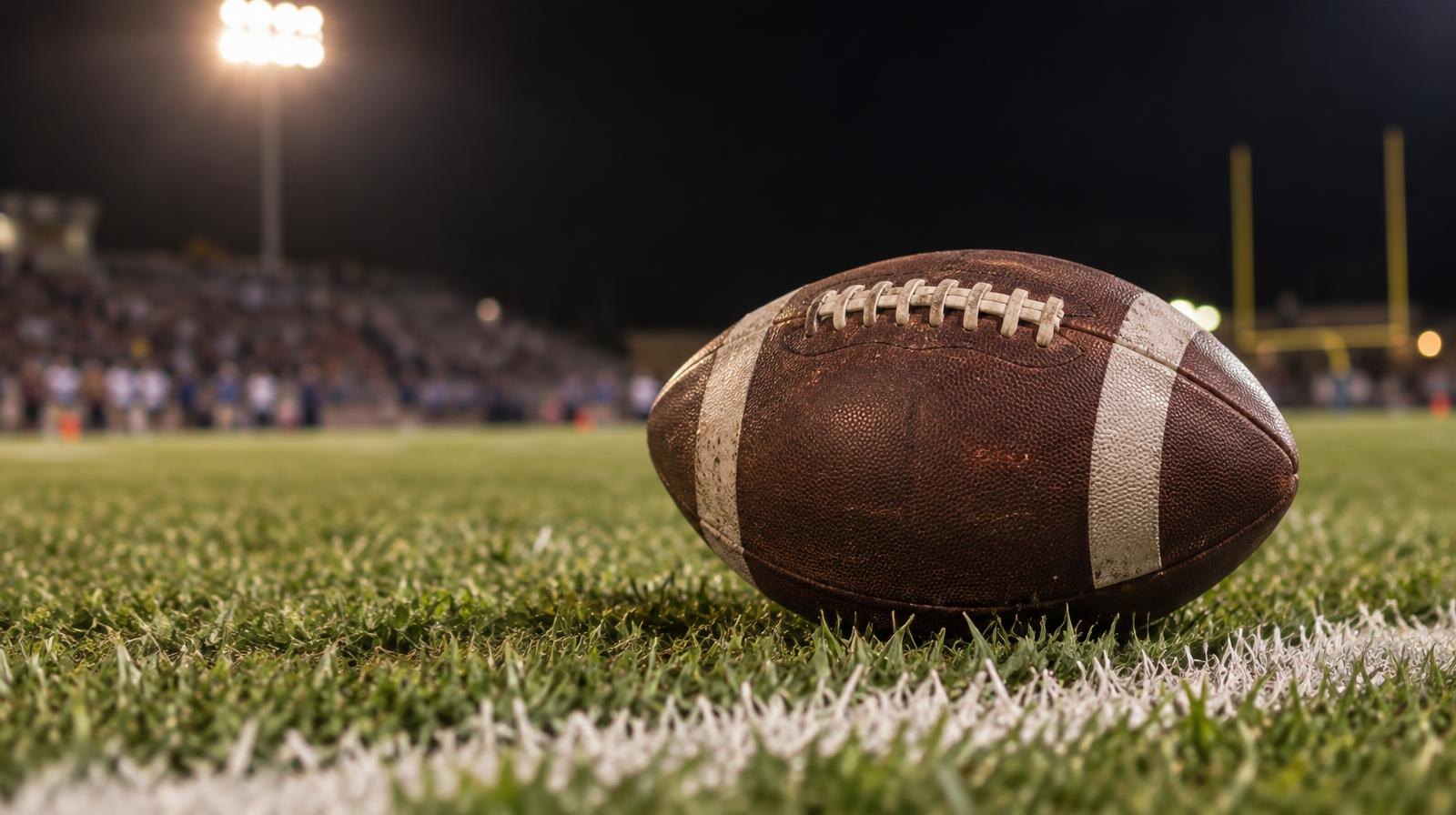An American football on green grass near a white yard line under bright stadium lights.
