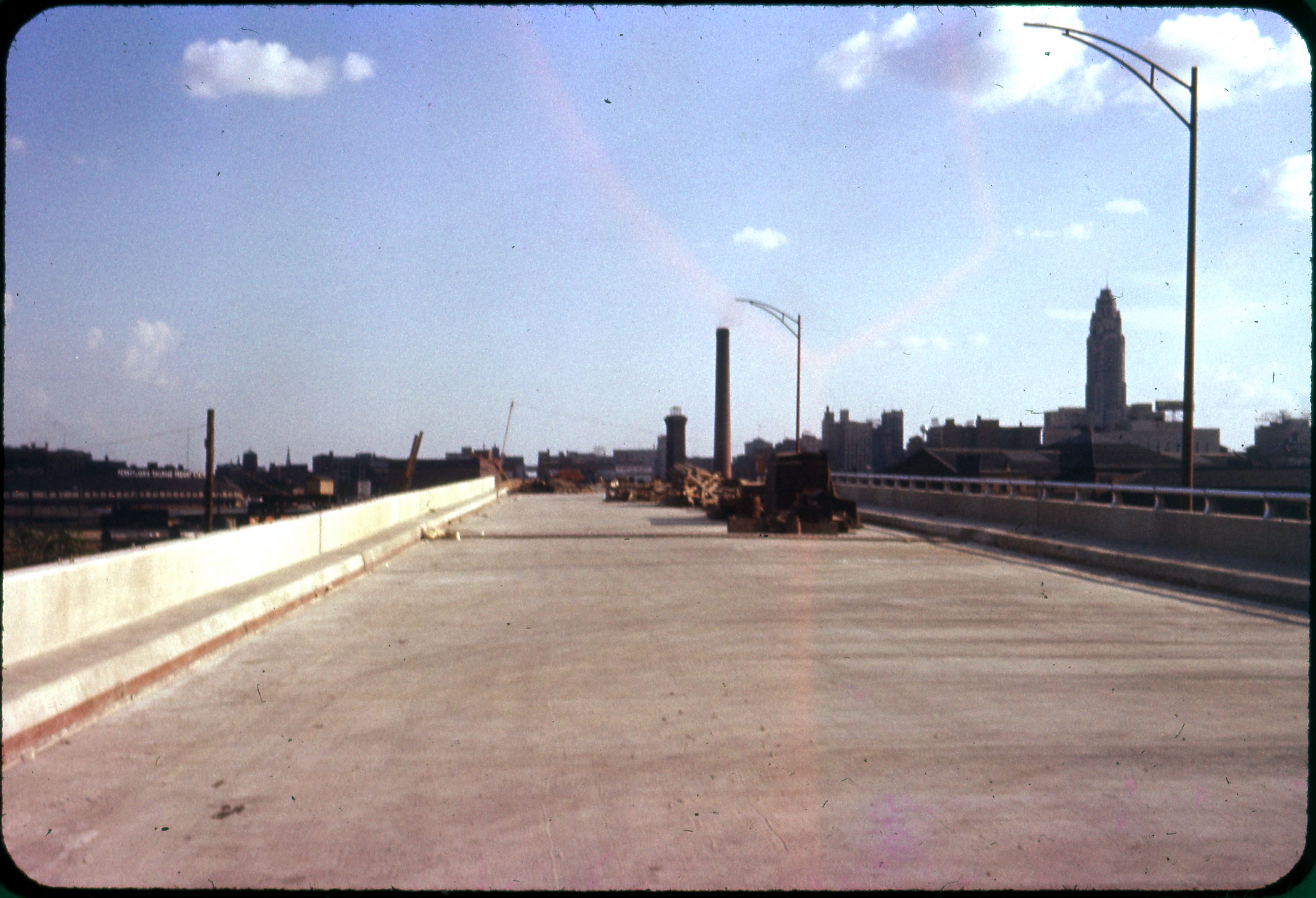Wide concrete viaduct deck under construction with equipment and a city skyline in the background.
