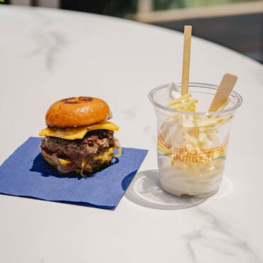 A smashburger with melted cheese next to a branded milkshake cup topped with French fries on a marble table.