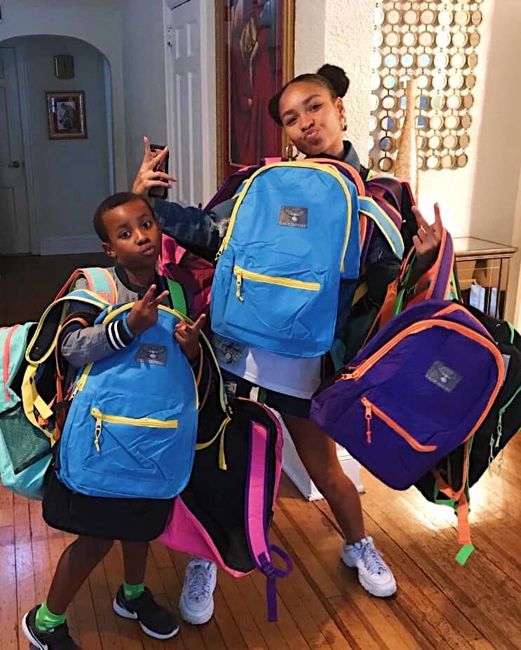 Two children make peace signs surrounded by a large pile of colorful donated backpacks.