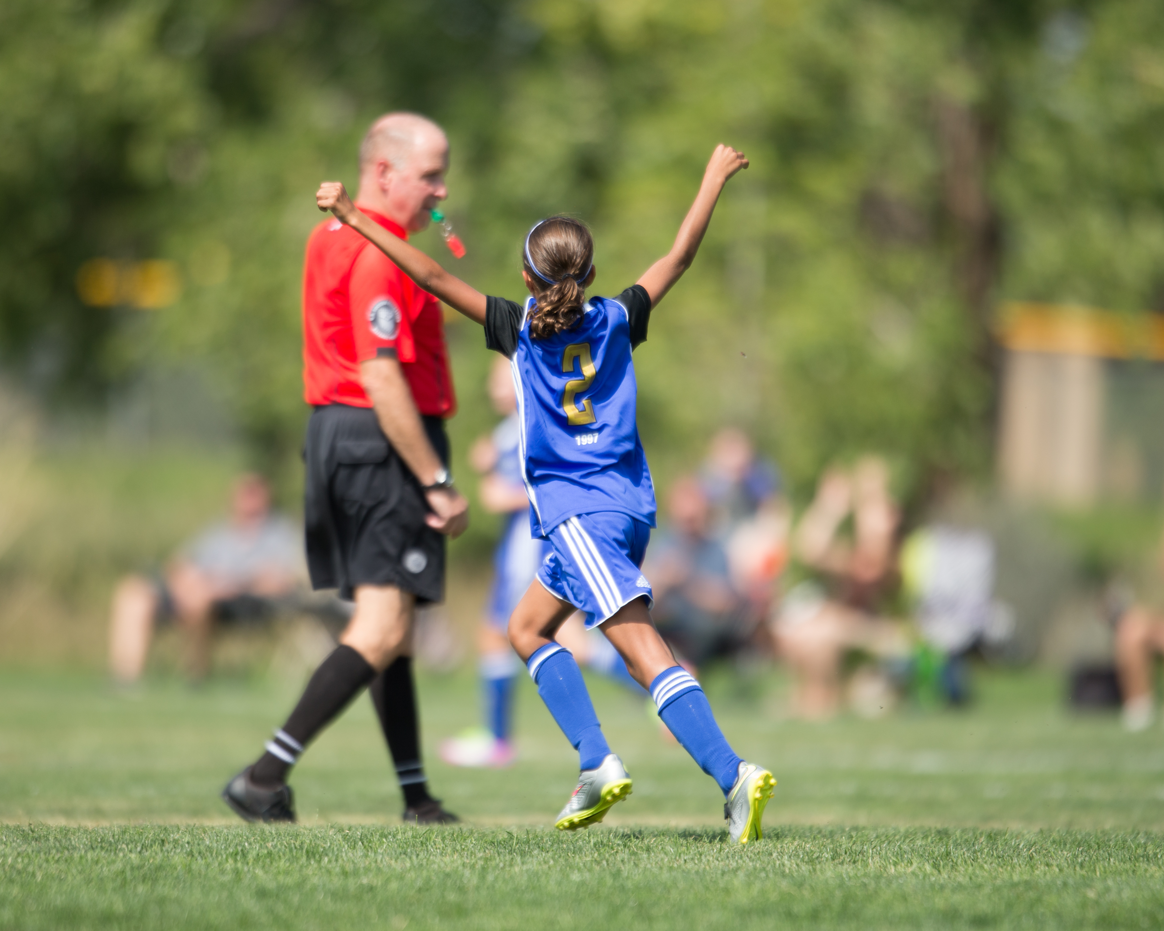 Youth soccer player in blue uniform raises both arms on a grass field.