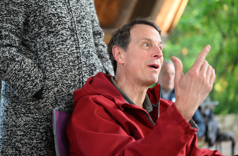 A man in a red jacket seated outdoors, gesturing with one finger raised while speaking.