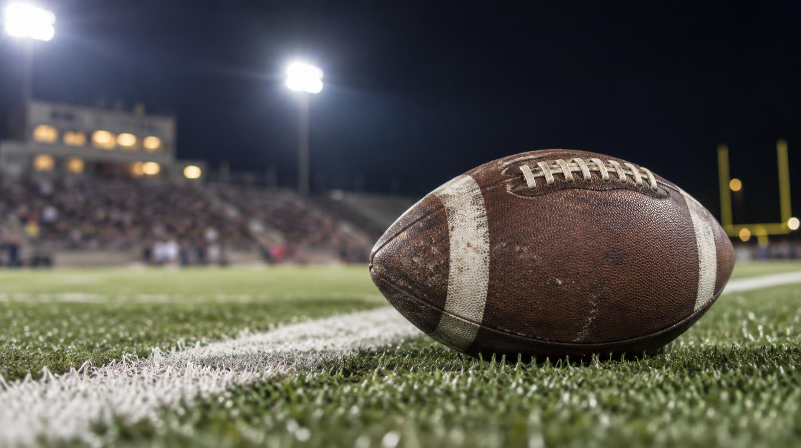 A brown football resting on green artificial turf beside a white yard line at night.