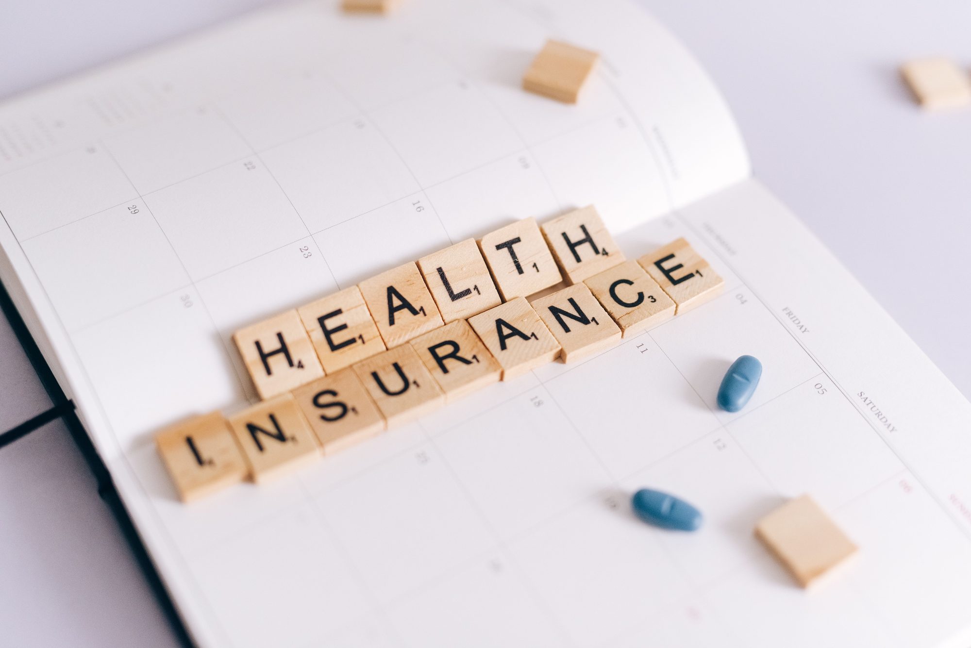 Close-up of health insurance spelled out with Scrabble tiles on a planner, alongside medication pills.