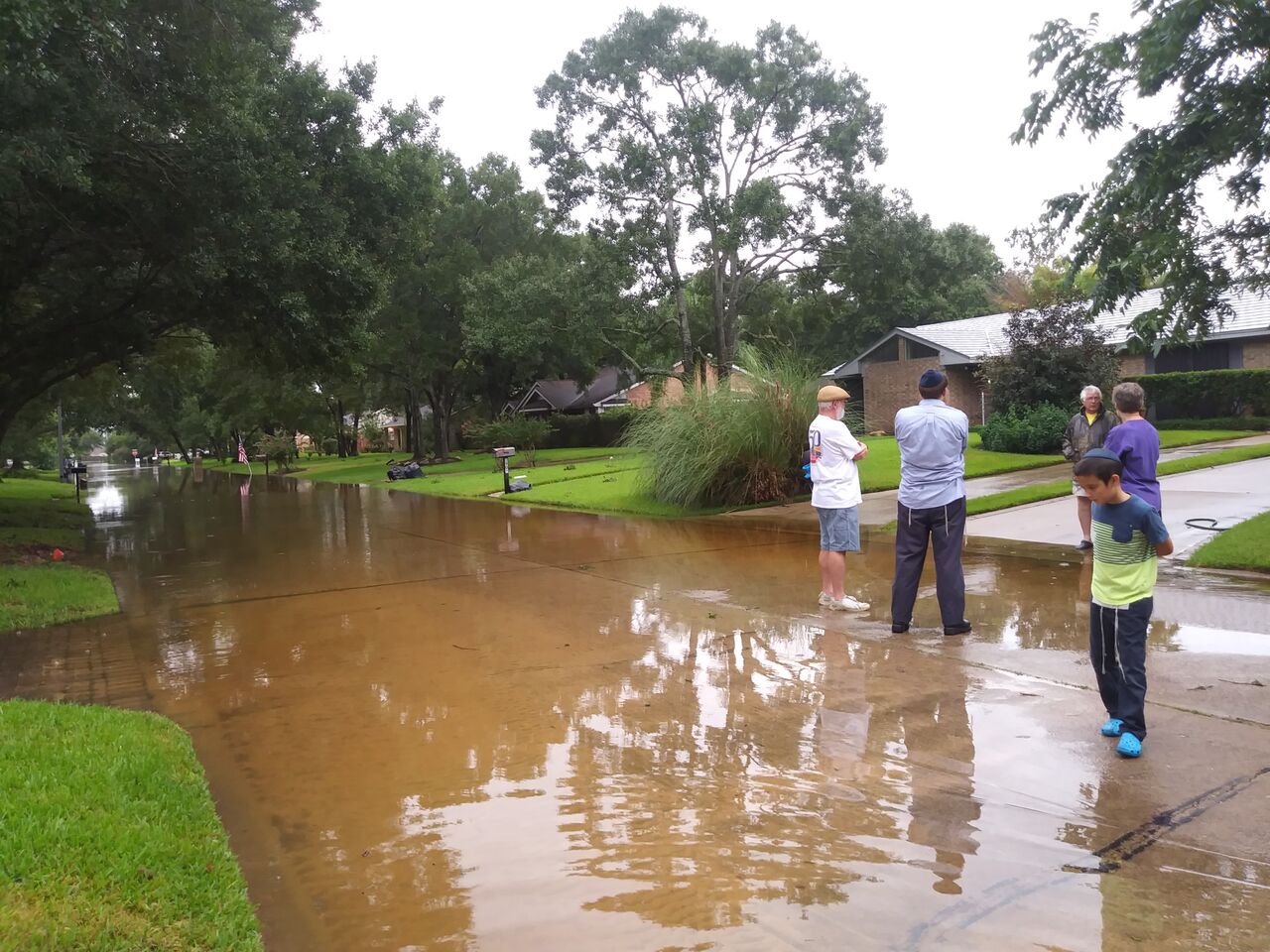 People stand beside a flooded suburban street covered in brown water.