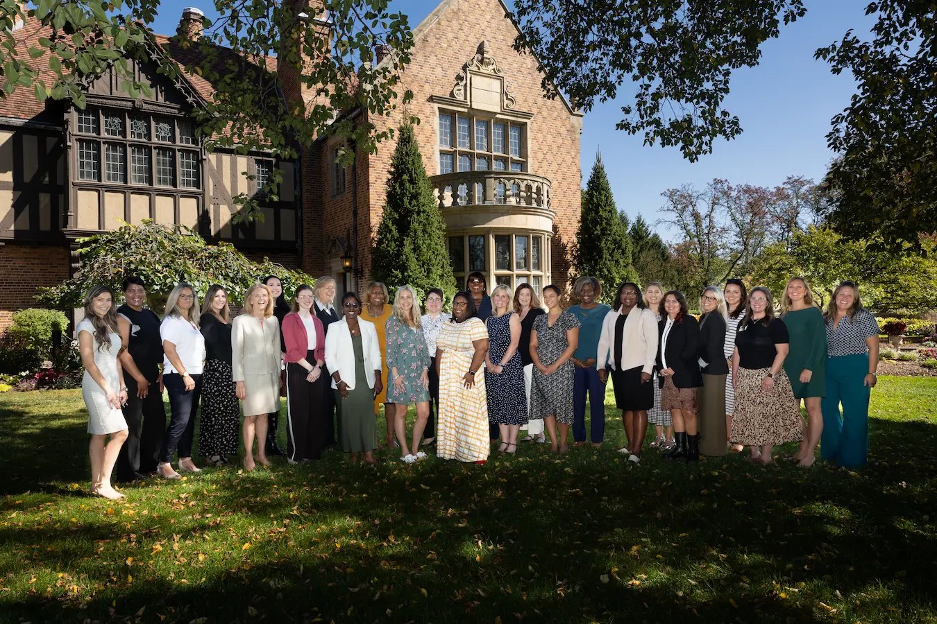Group of women in professional attire posed on a lawn in front of a Tudor-revival brick estate.