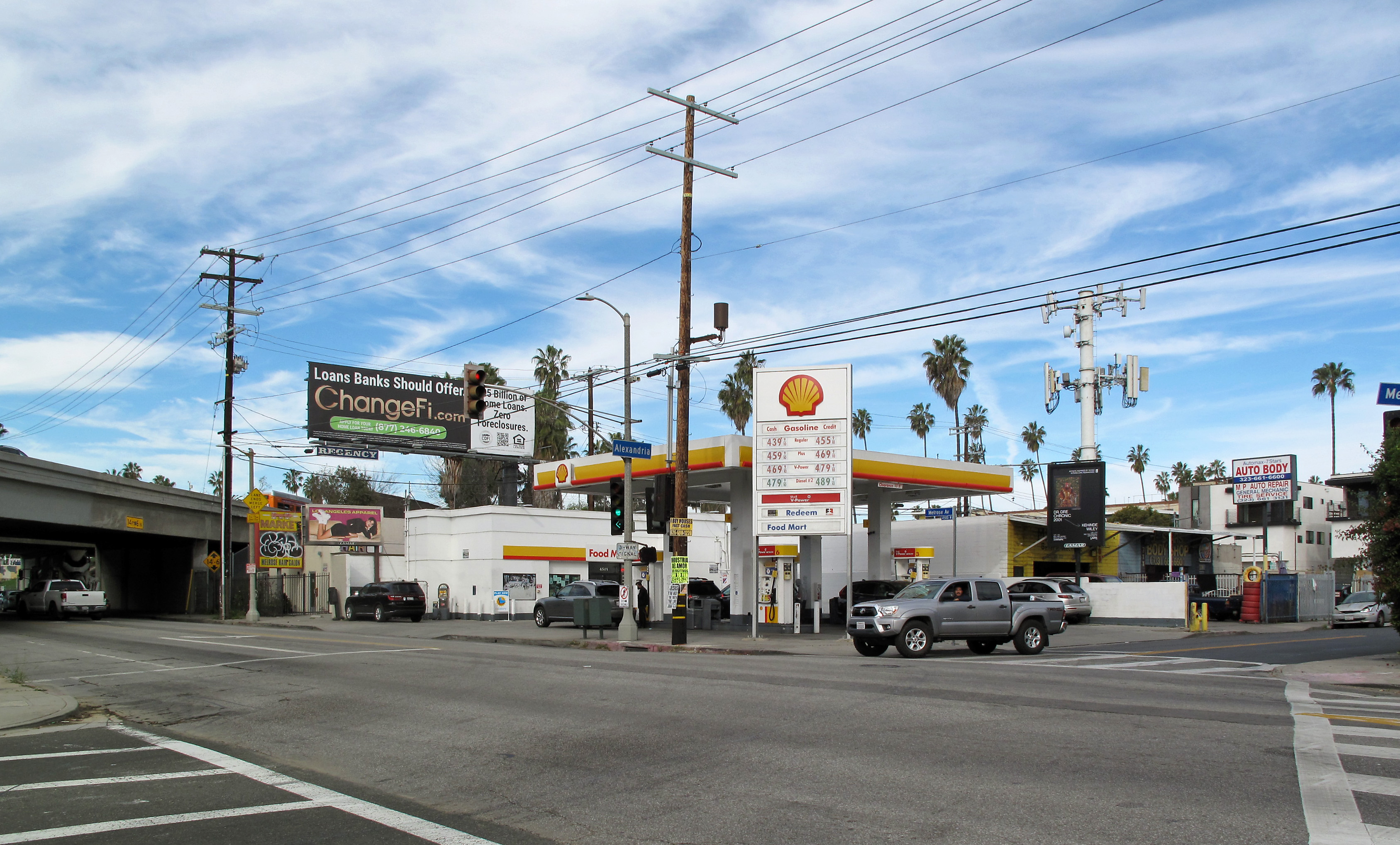 Shell gas station with price signs at a Los Angeles intersection, freeway overpass behind.