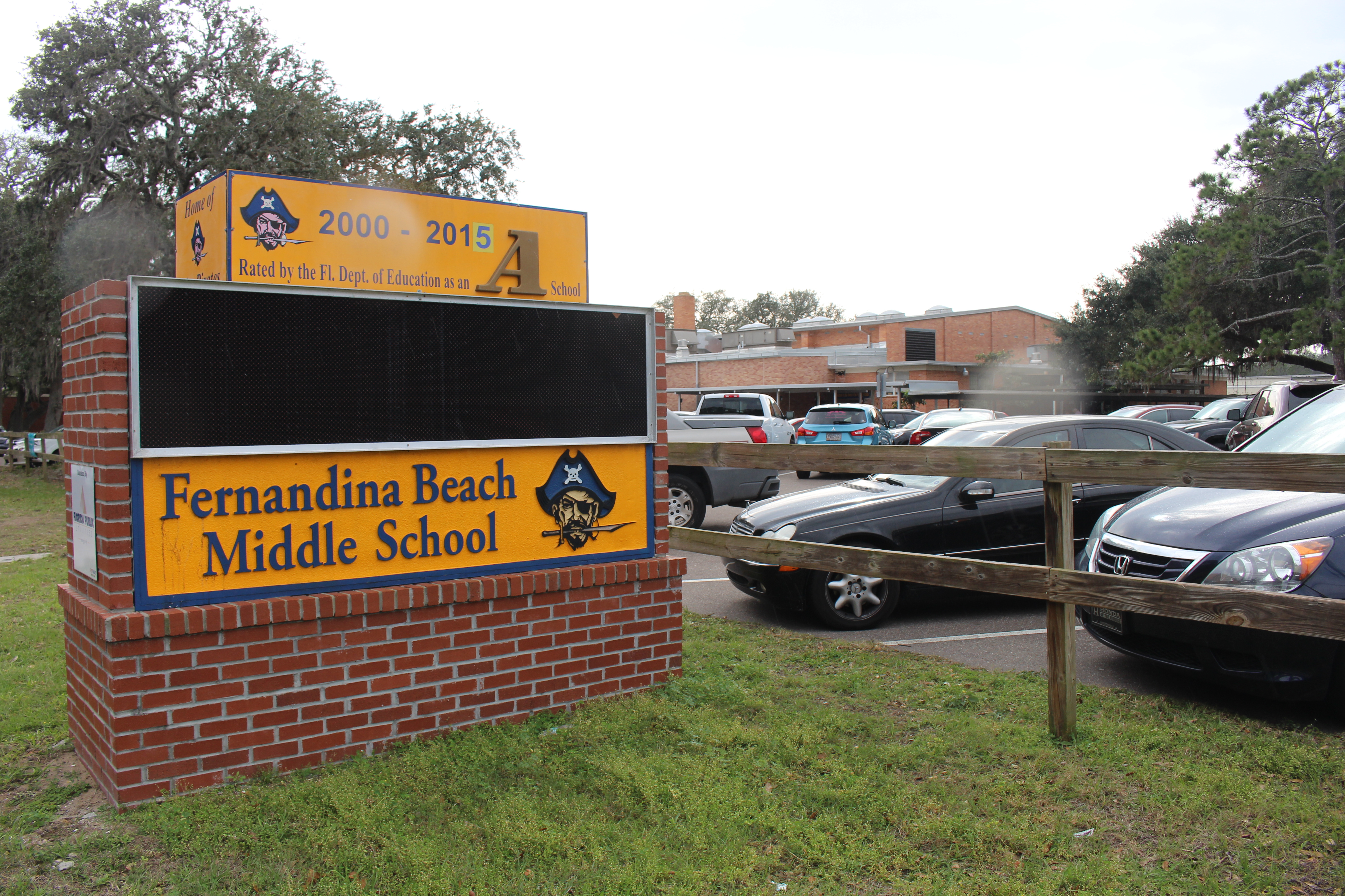 School entrance sign on a brick base with a pirate mascot logo and parking lot behind.