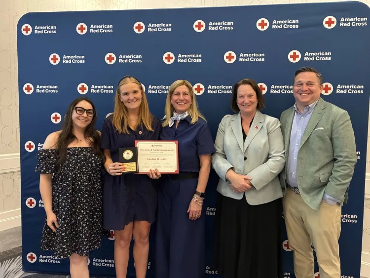 Five people pose in front of a Red Cross step-and-repeat backdrop; one holds a framed certificate and plaque.
