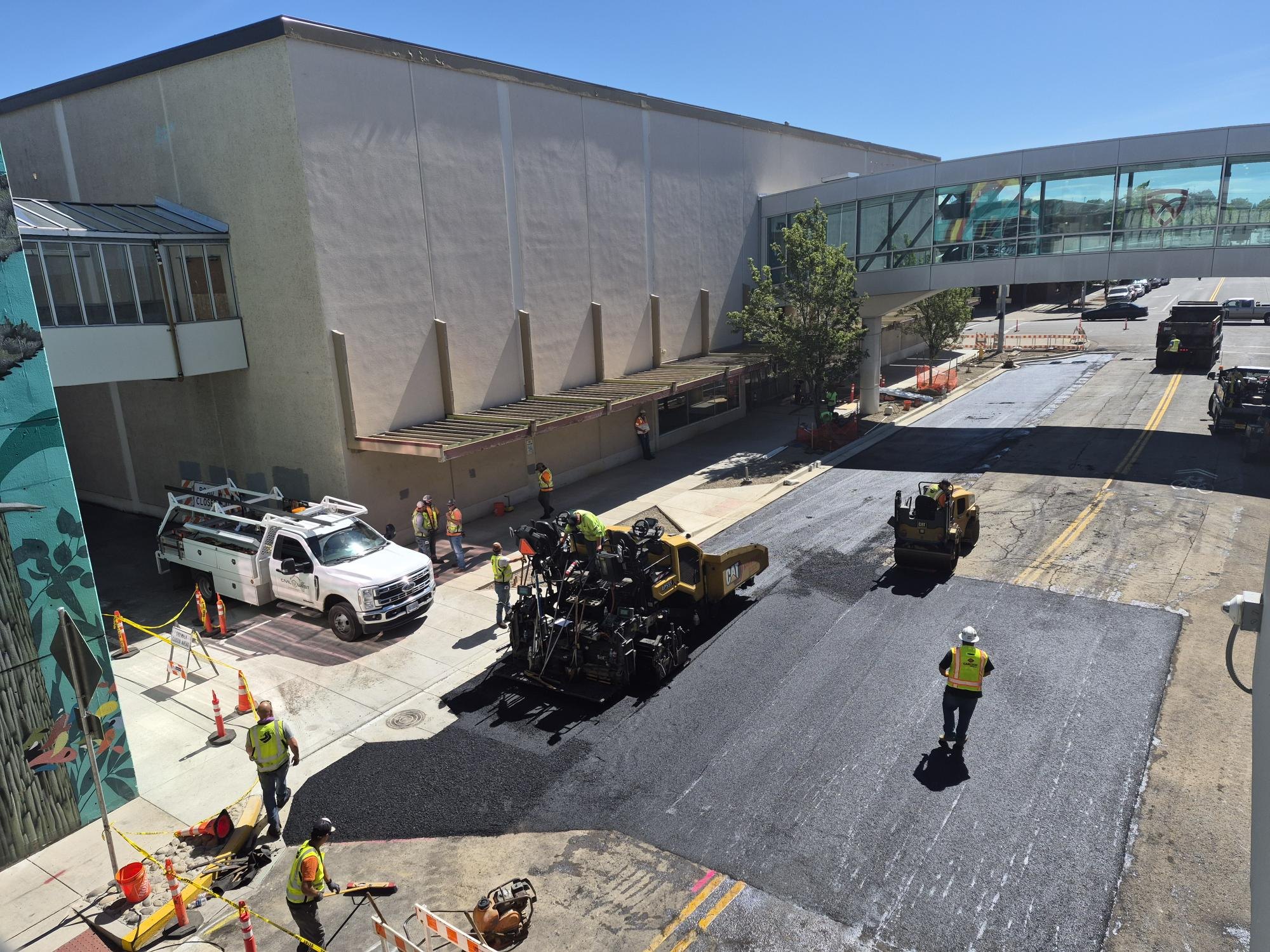 Aerial view of road paving operation with workers in high-visibility vests and heavy equipment between buildings.