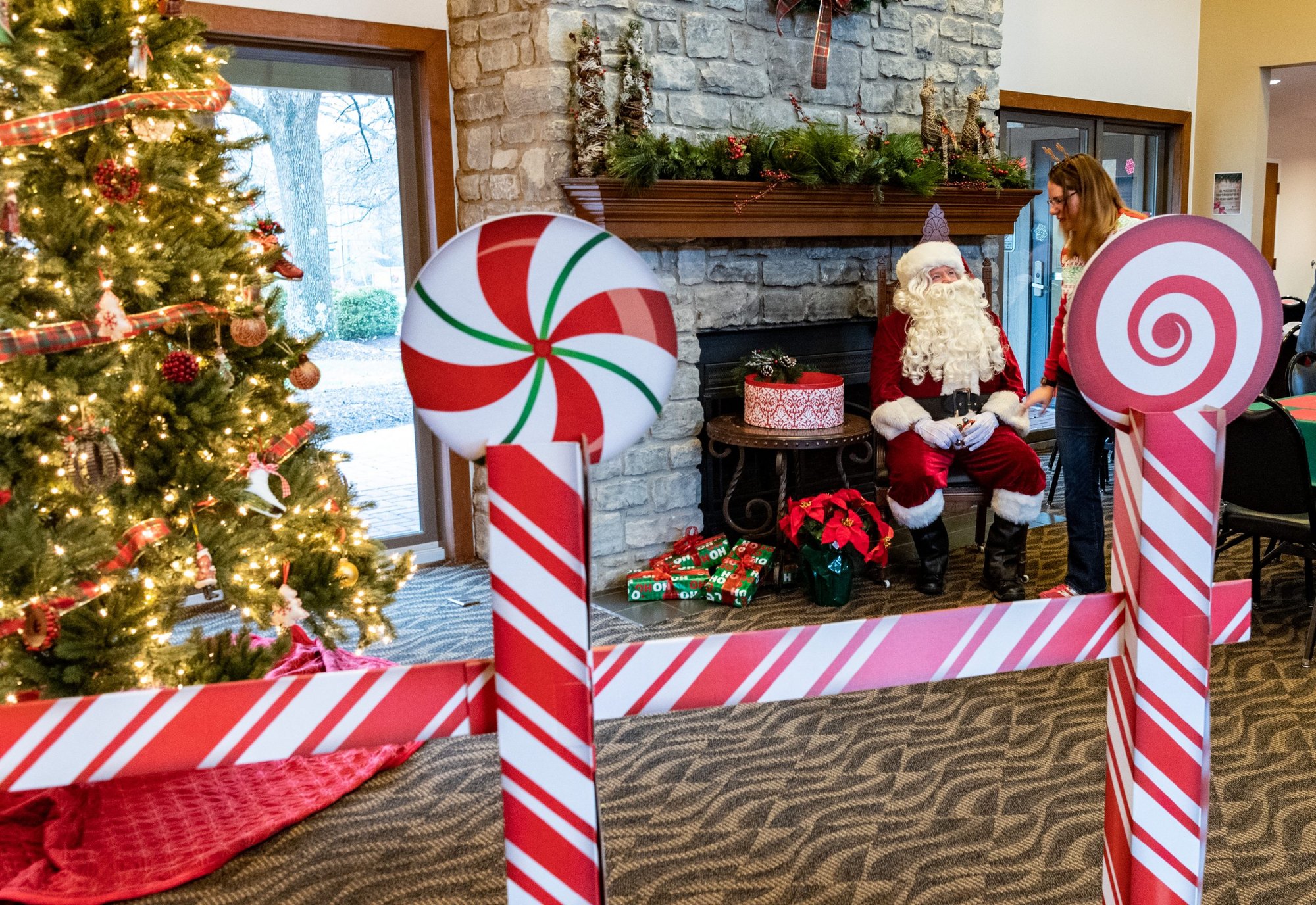 Person dressed as Santa Claus seated near a decorated stone fireplace with Christmas tree and holiday props.