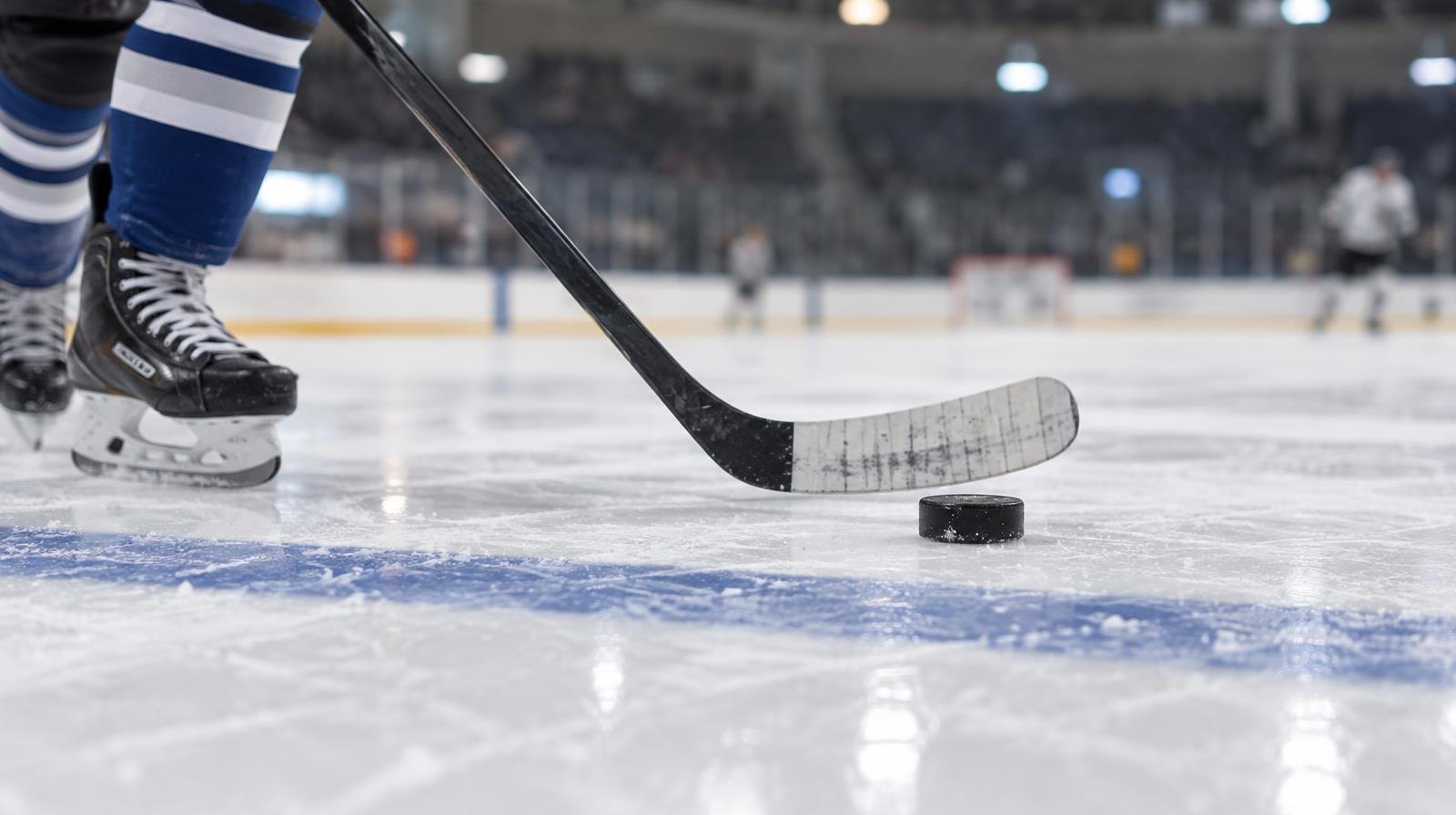 A hockey stick and puck on the ice near a blue line in an indoor rink.