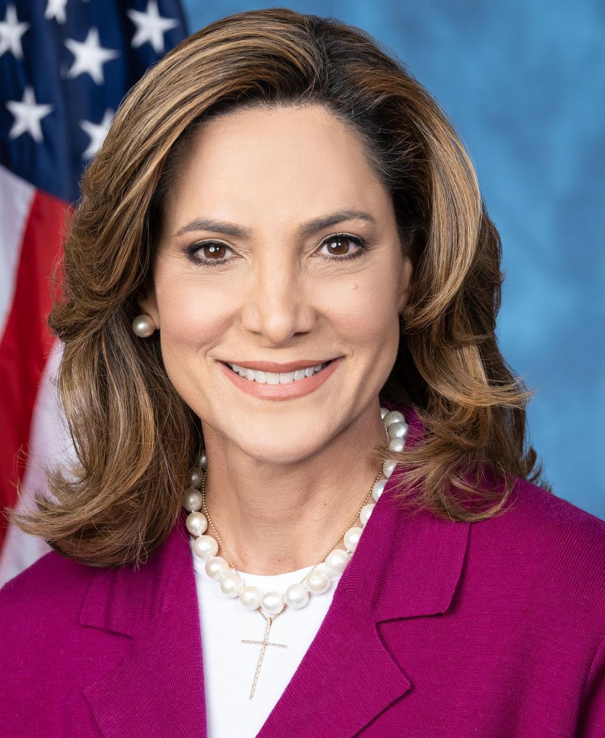 Woman in a magenta blazer with pearl necklace posed before an American flag.