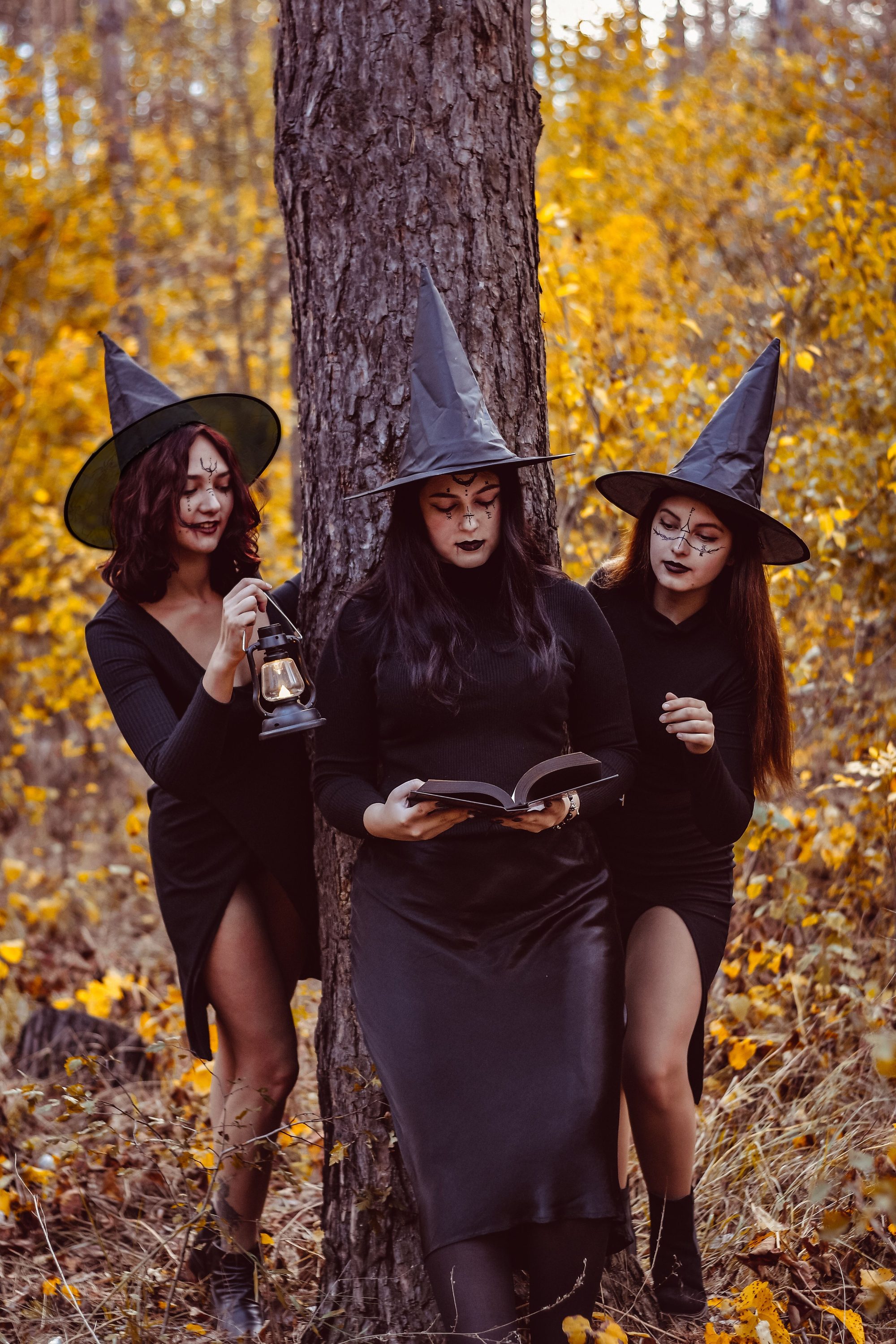 Three women in witch costumes in an autumn forest reading a spellbook.