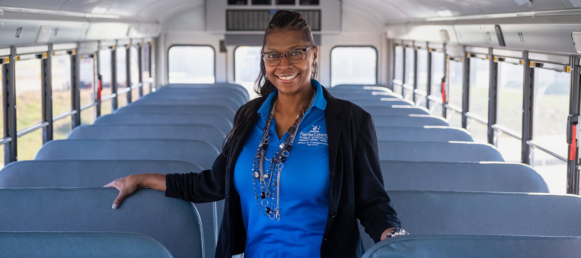 A woman in a blue polo shirt stands smiling inside an empty school bus.
