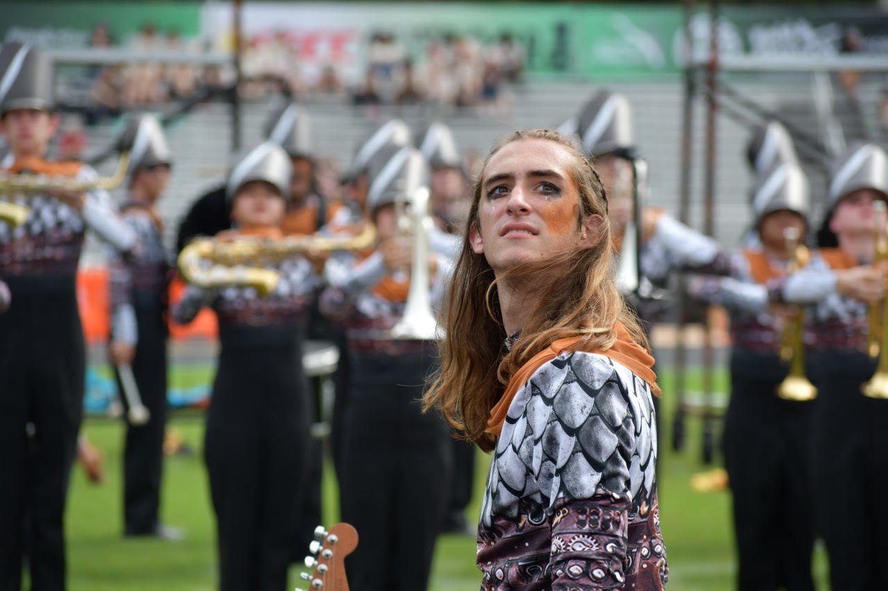 A costumed performer stands on a grass field with uniformed marching band members holding brass instruments behind her.