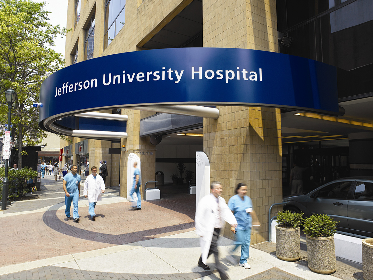Hospital entrance with blue curved sign reading Jefferson University Hospital and people in scrubs walking outside.