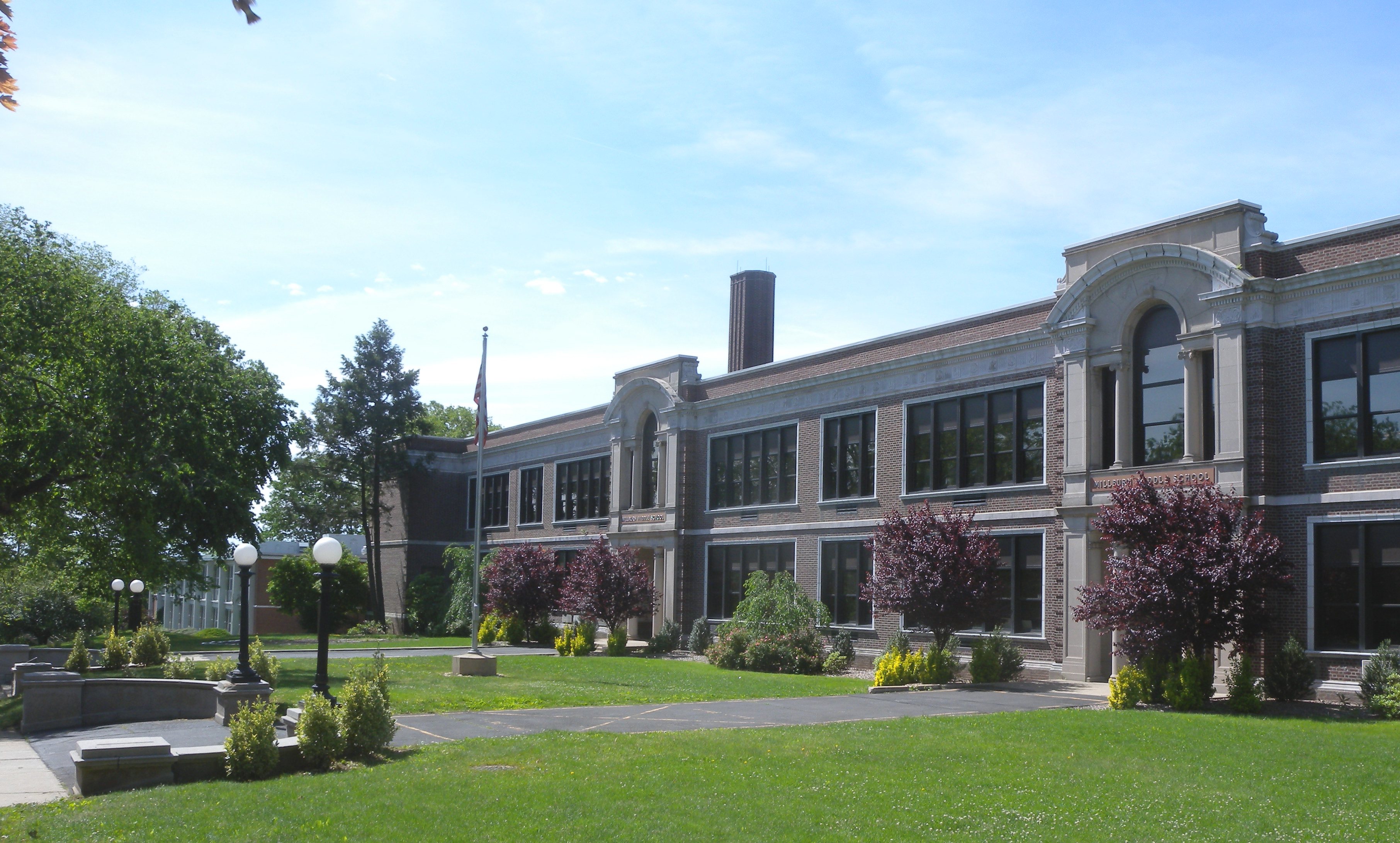 Brick and stone school building with arched windows, ornamental trees, and a green lawn on a sunny day.