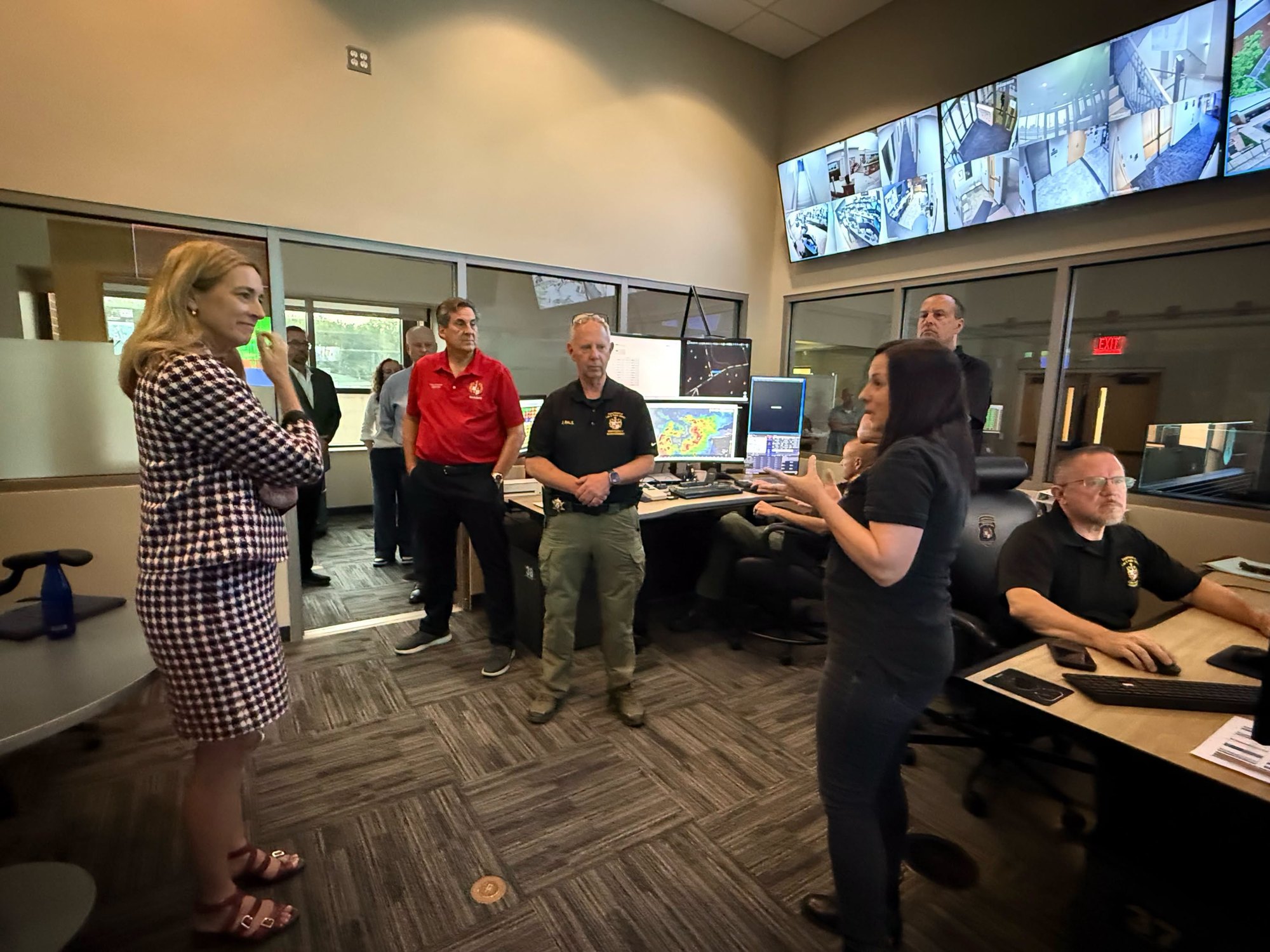 Officials gathered in an emergency communications center with large monitors on the wall showing weather maps and surveillance feeds.