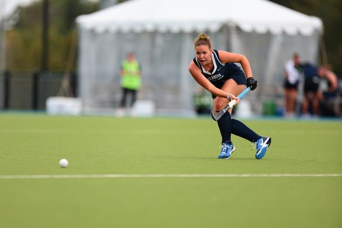 Female field hockey player in navy uniform dribbling a ball on green turf.