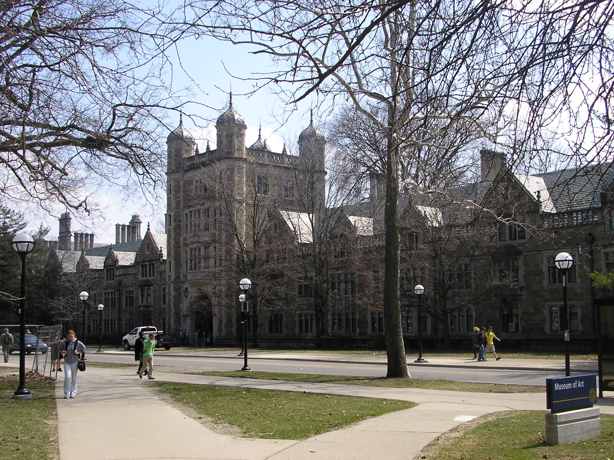 Gothic stone university buildings with bare trees and pedestrians on campus walkways.
