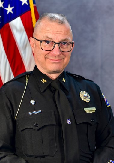 Police officer in black uniform with gold badge and command stars, posed before an American flag.