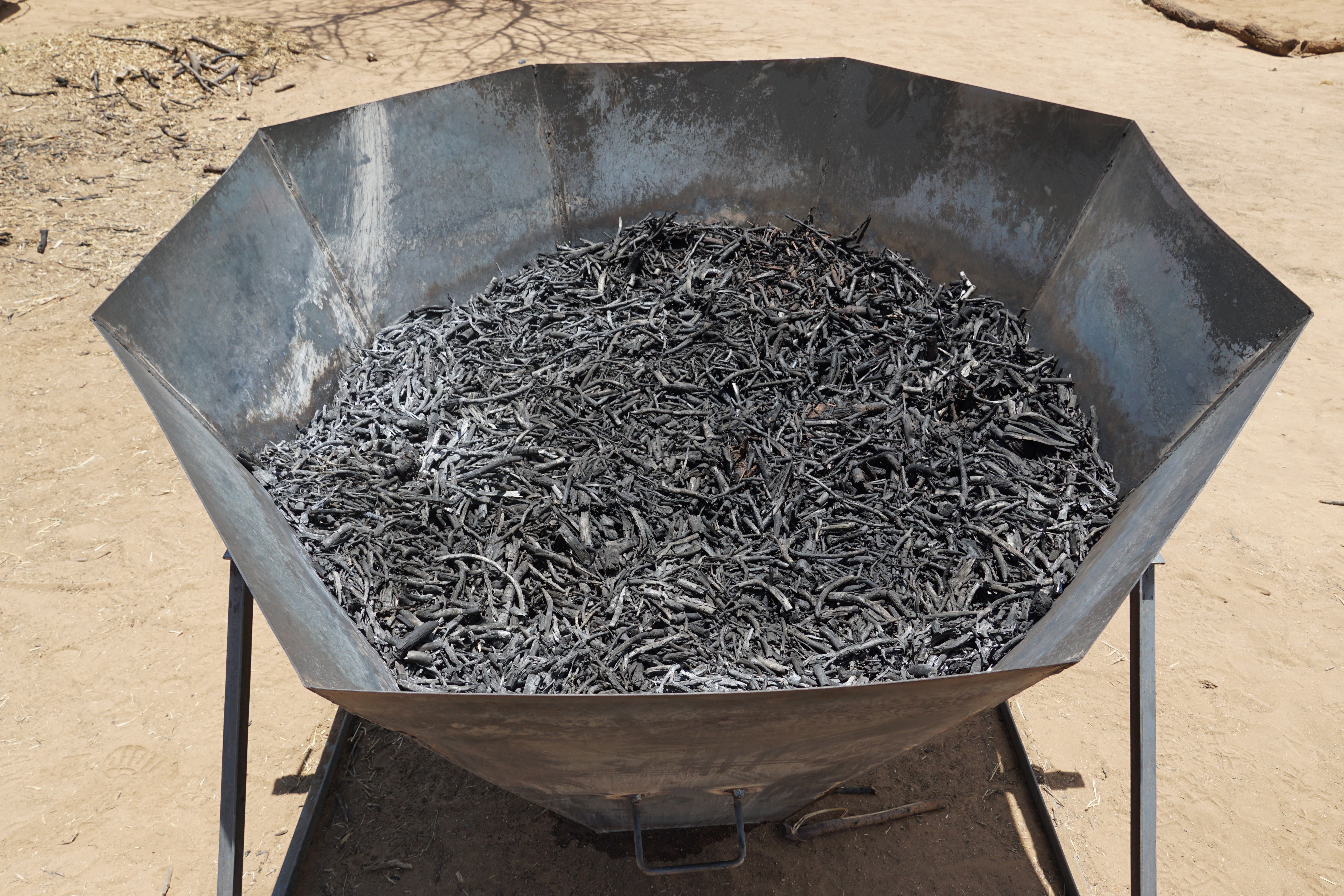 Large octagonal metal kiln filled with dark charred biomass on a metal stand outdoors.