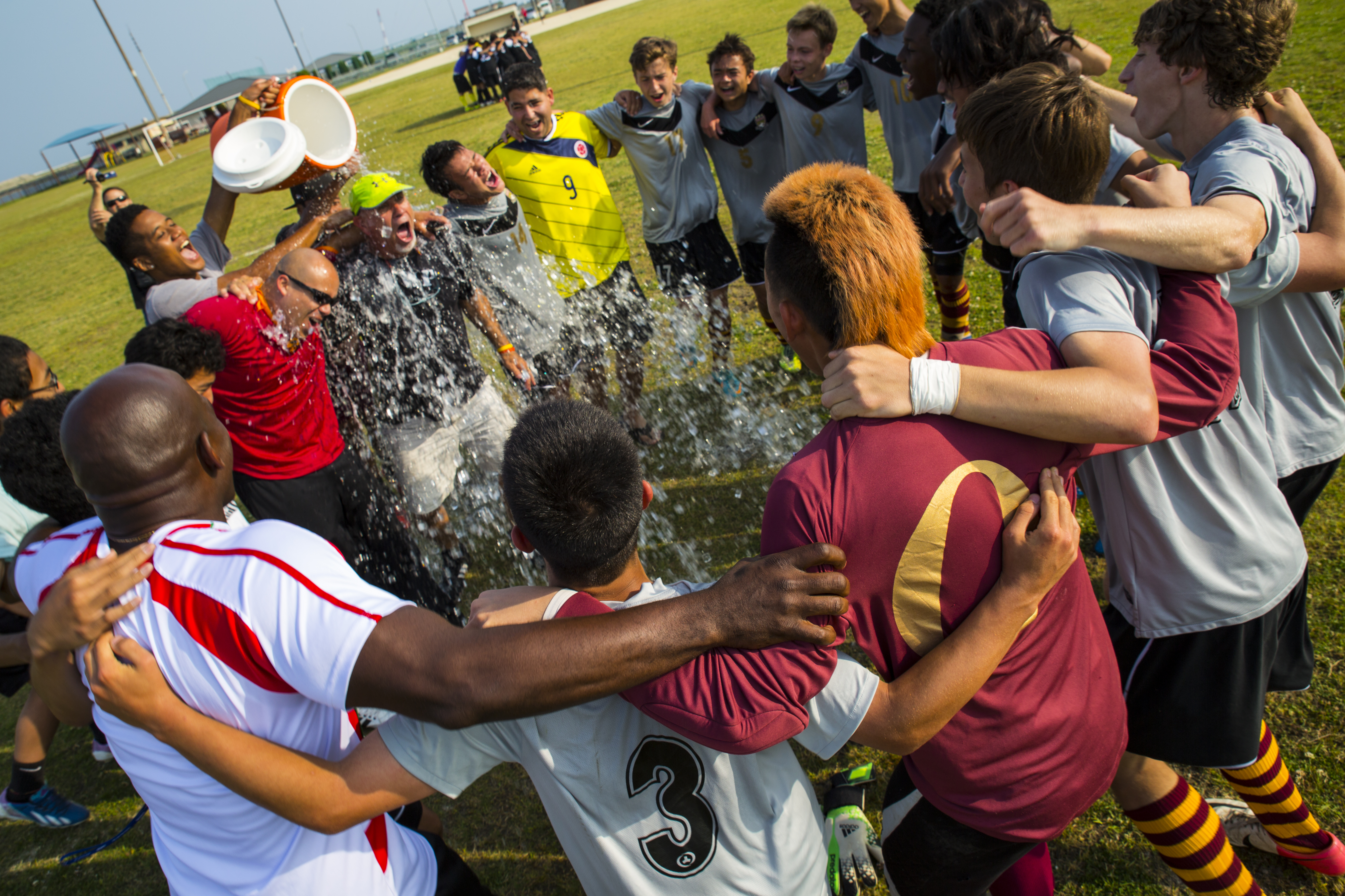 Soccer players dump a cooler of water on their coach on a grass field after winning a championship.