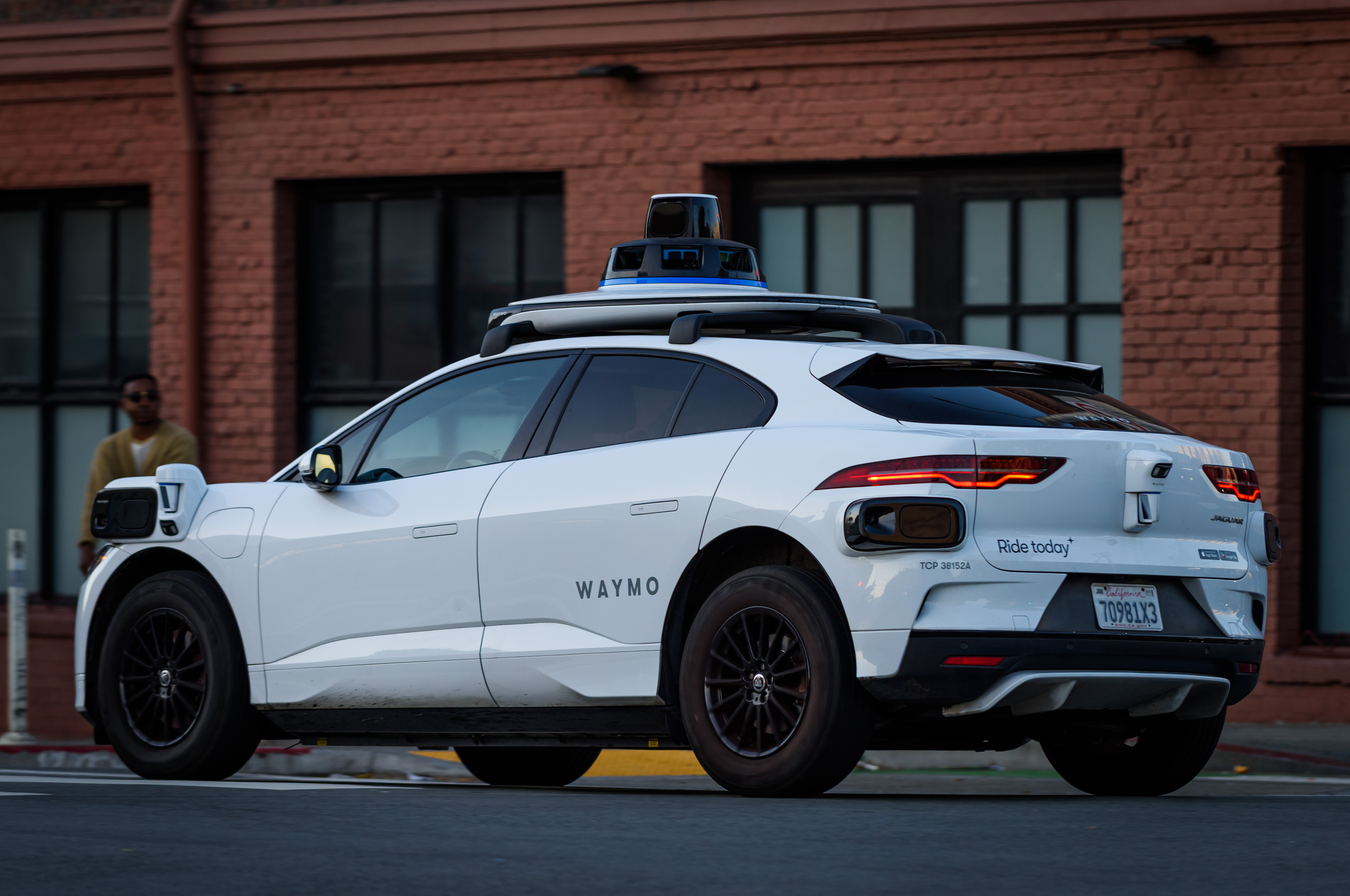 Rear view of a white Waymo-branded autonomous vehicle with roof sensors on a city street.