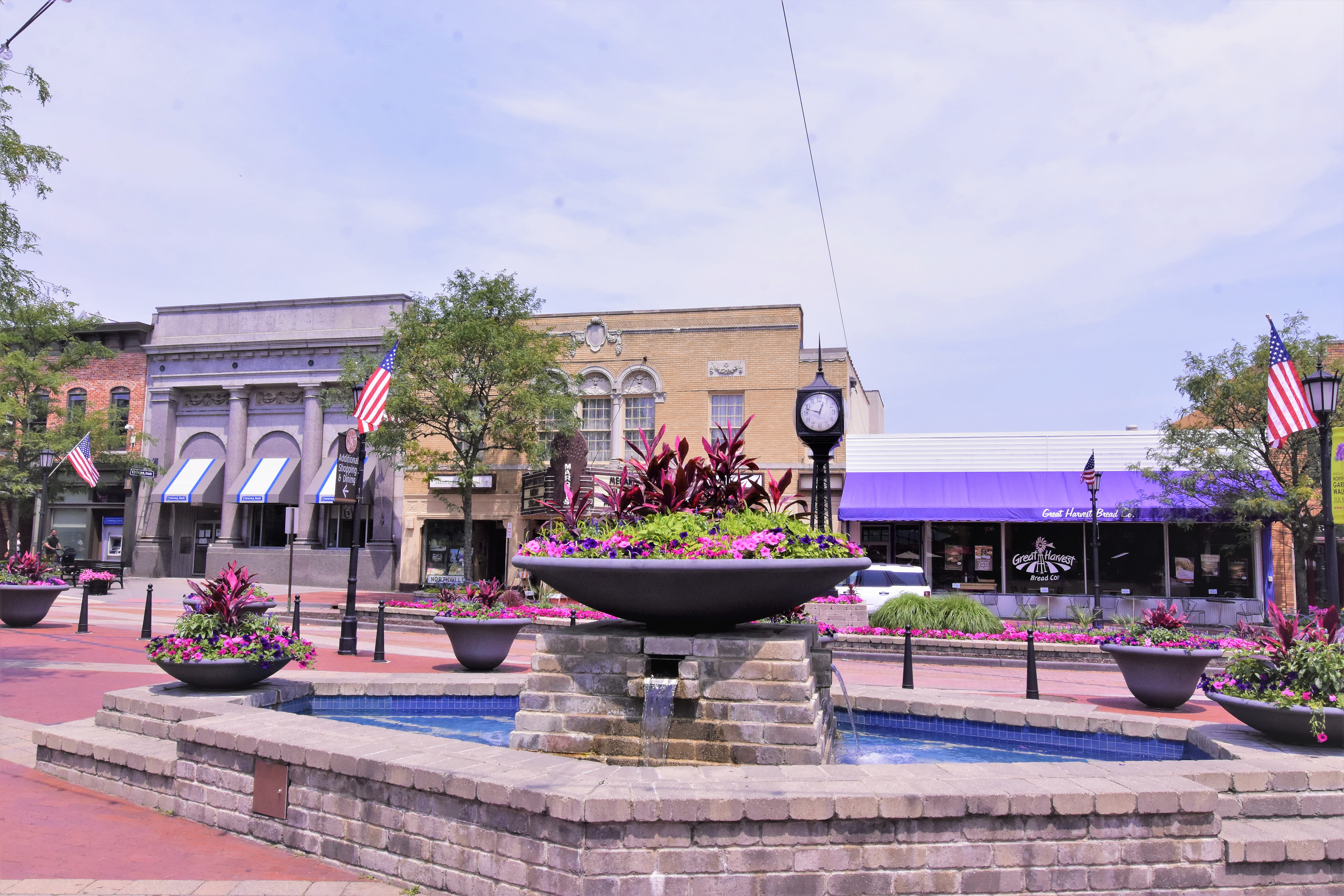 Brick town square with a tiered fountain, flower planters, and historic storefronts.