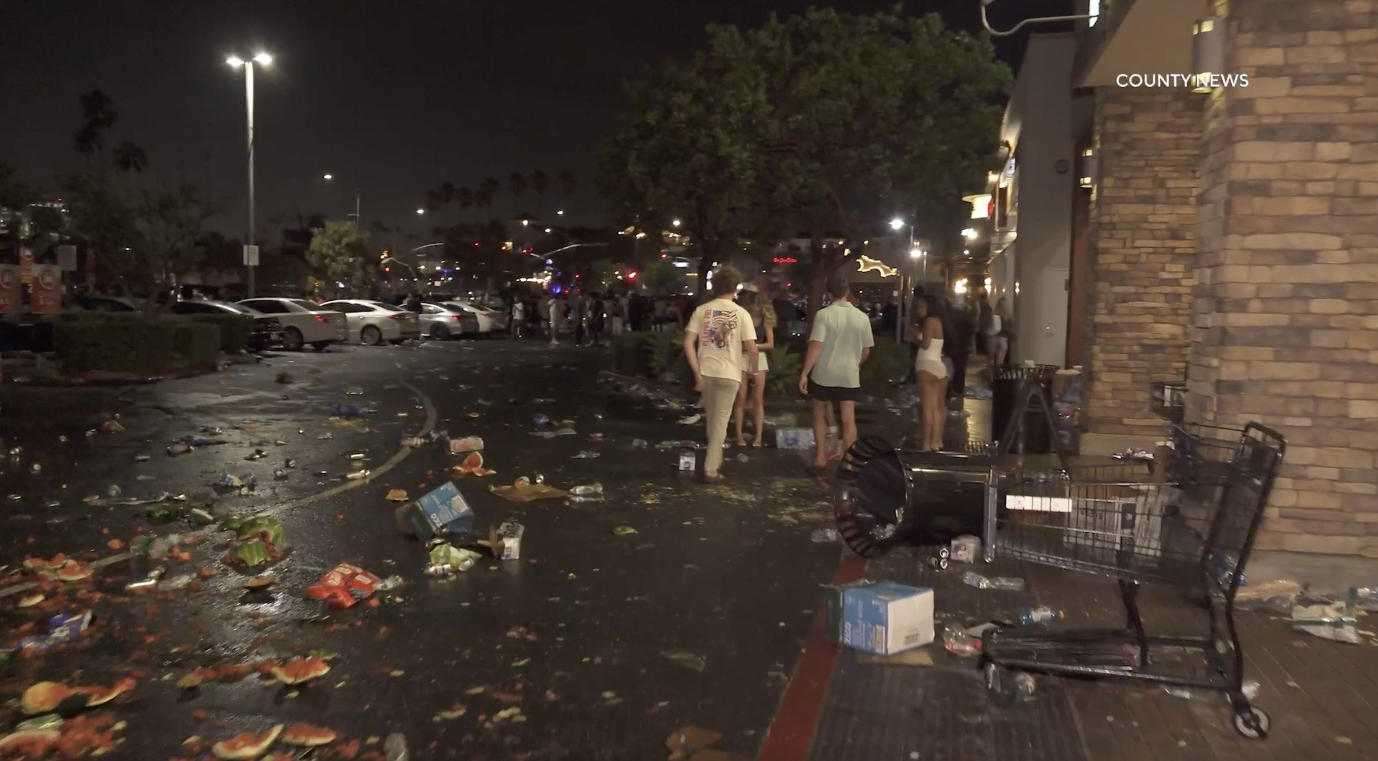 Nighttime scene of debris scattered on wet ground outside a supermarket entrance with people walking through the area.