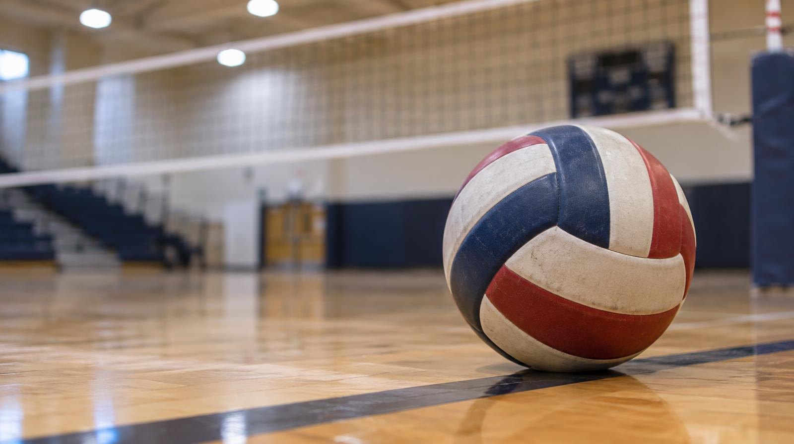 A volleyball sits on a hardwood gym floor near the net under bright overhead lights.