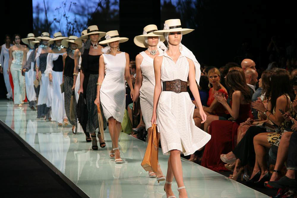 A line of models in white dresses and straw hats walks a reflective runway before a seated audience.