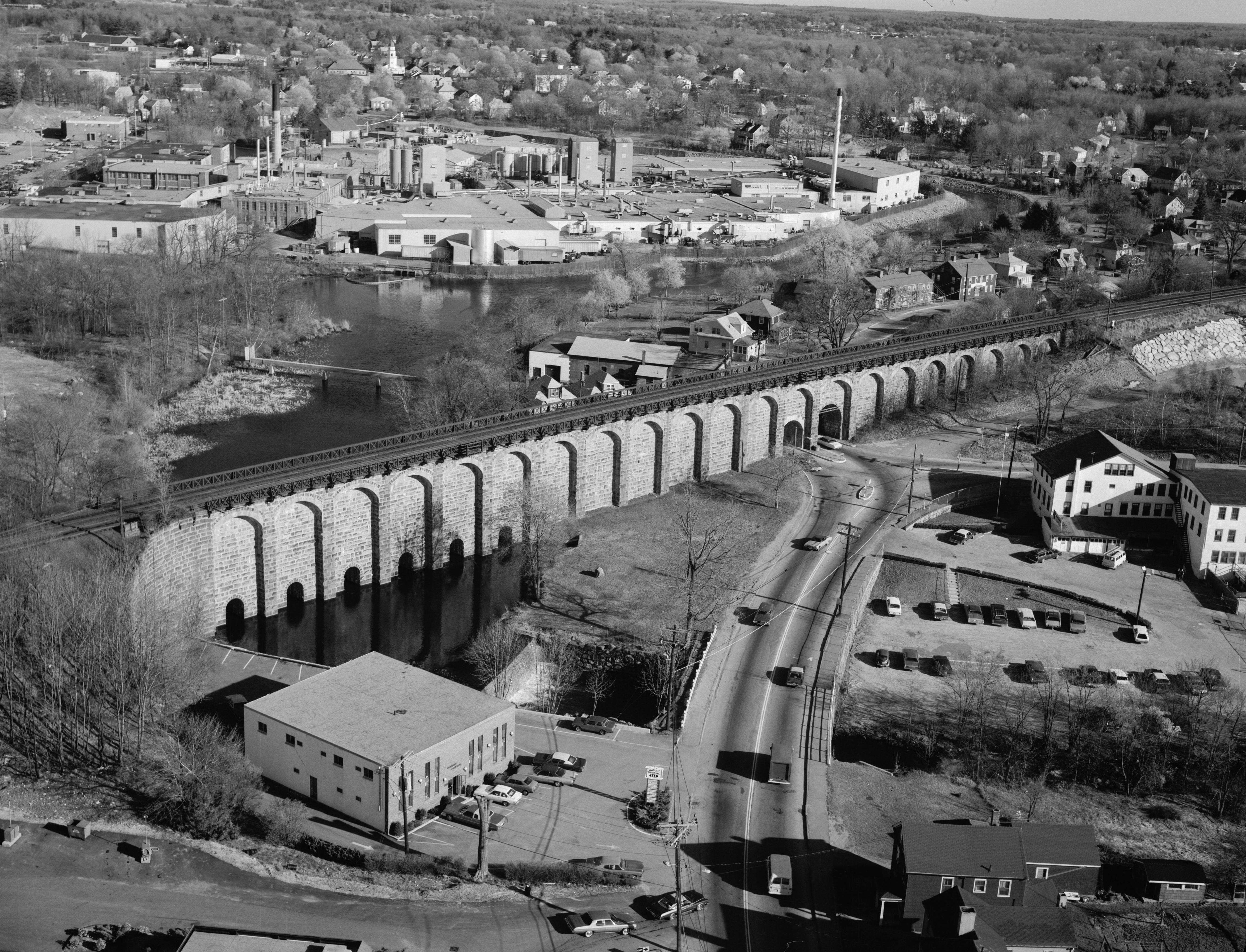 Aerial black-and-white photo of a large stone arch railroad viaduct with industrial buildings behind it.