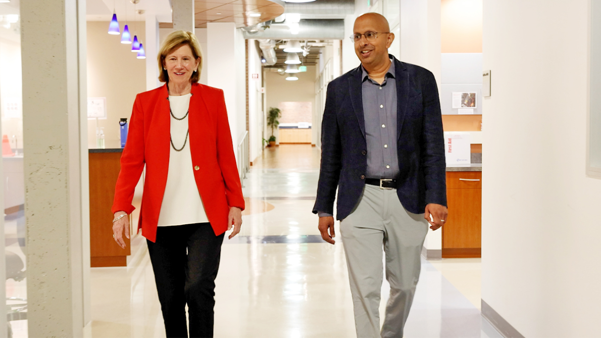 A woman in a red blazer and a man in a dark blazer walking side by side down a brightly lit institutional hallway.